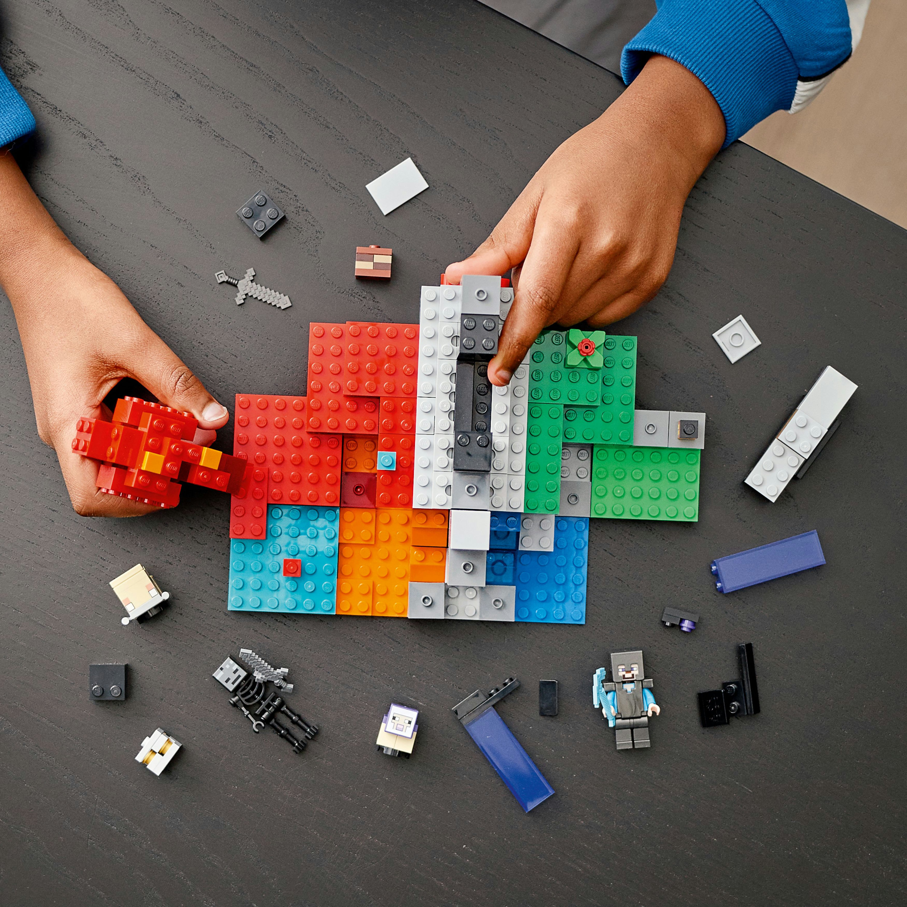 Hände bauen mit bunten LEGO-Steinen. Verschiedene LEGO-Figuren und Objekte sind um die Bausteinstruktur verstreut.
