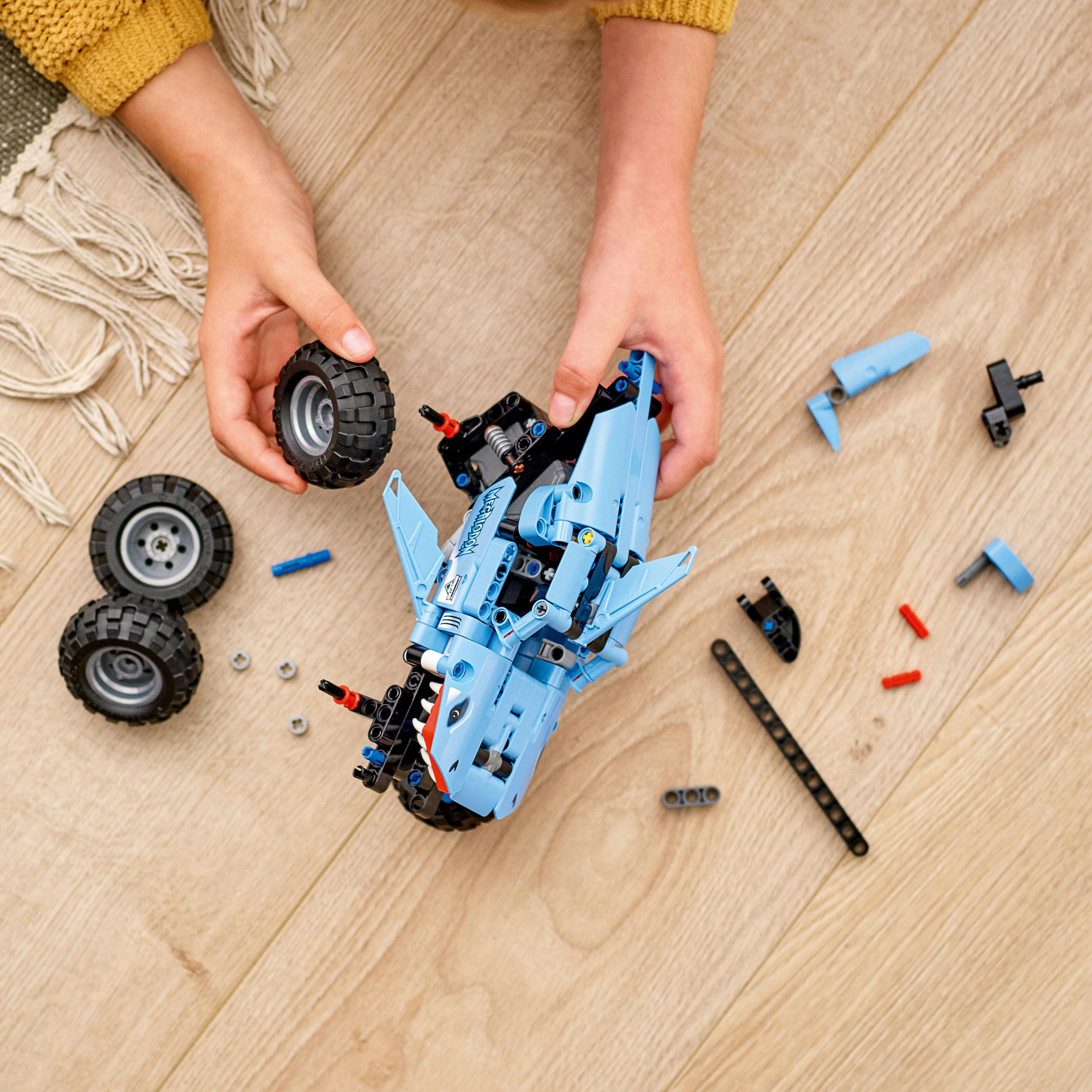 Hände halten ein blaues LEGO-Modell in Hai-Form, mit Rädern und anderen Teilen verstreut.
