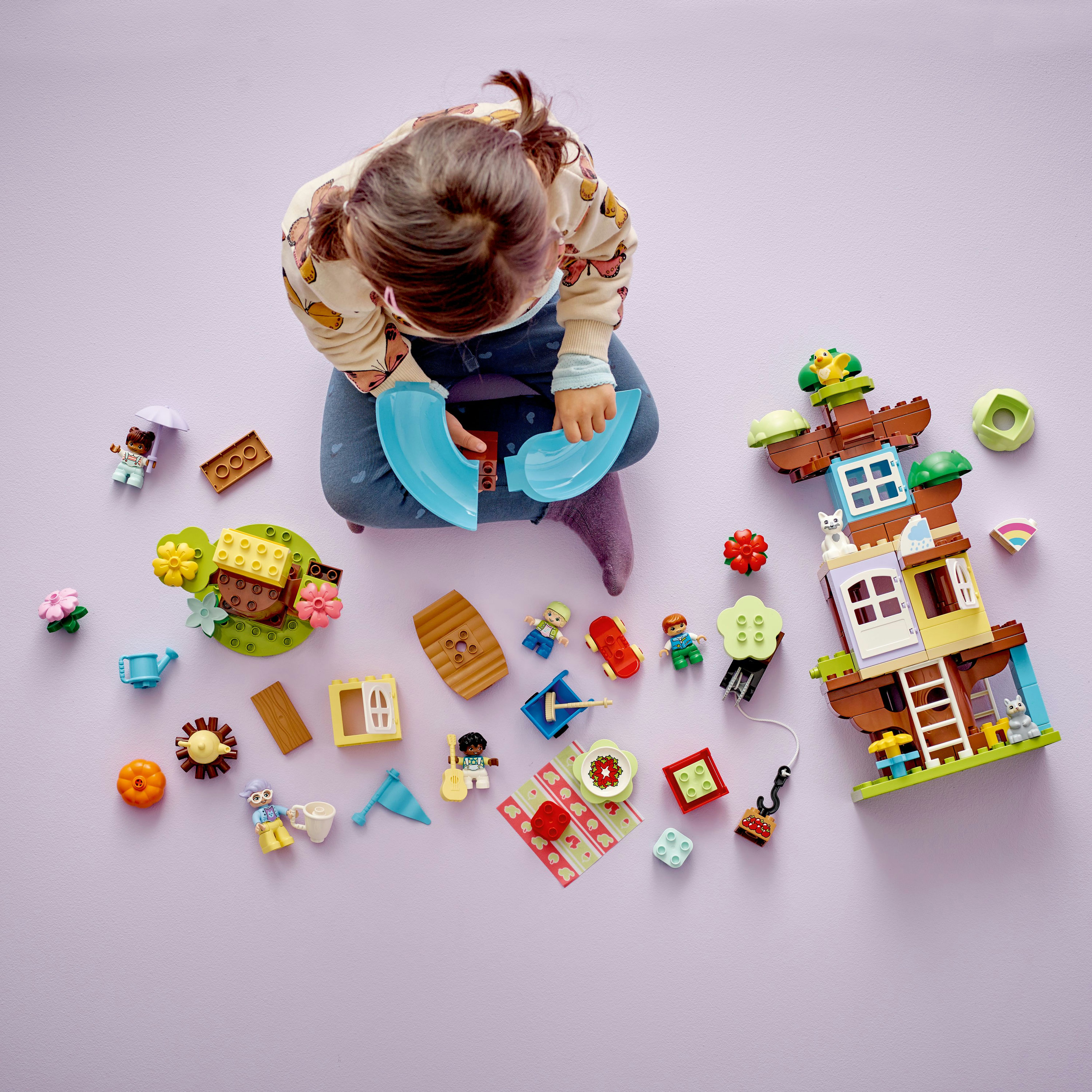 Ein Kind spielt mit Lego-Steinen, darunter ein Haus, Figuren und Zubehör, alles auf einer lila Oberfläche.