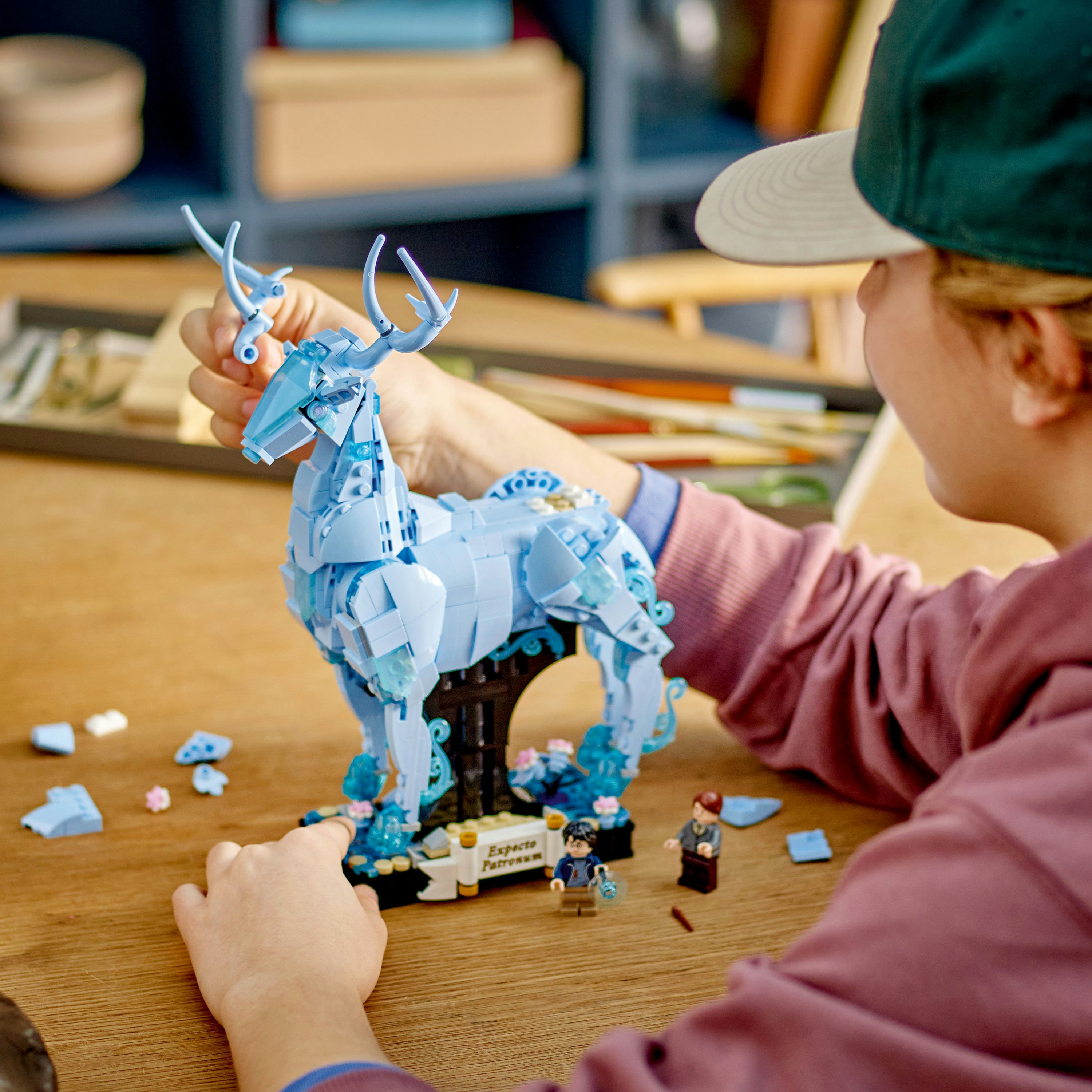 Ein Kind hält ein LEGO Patronus Set. Ein hellblaues Reh, Minifiguren und ein Holztisch.