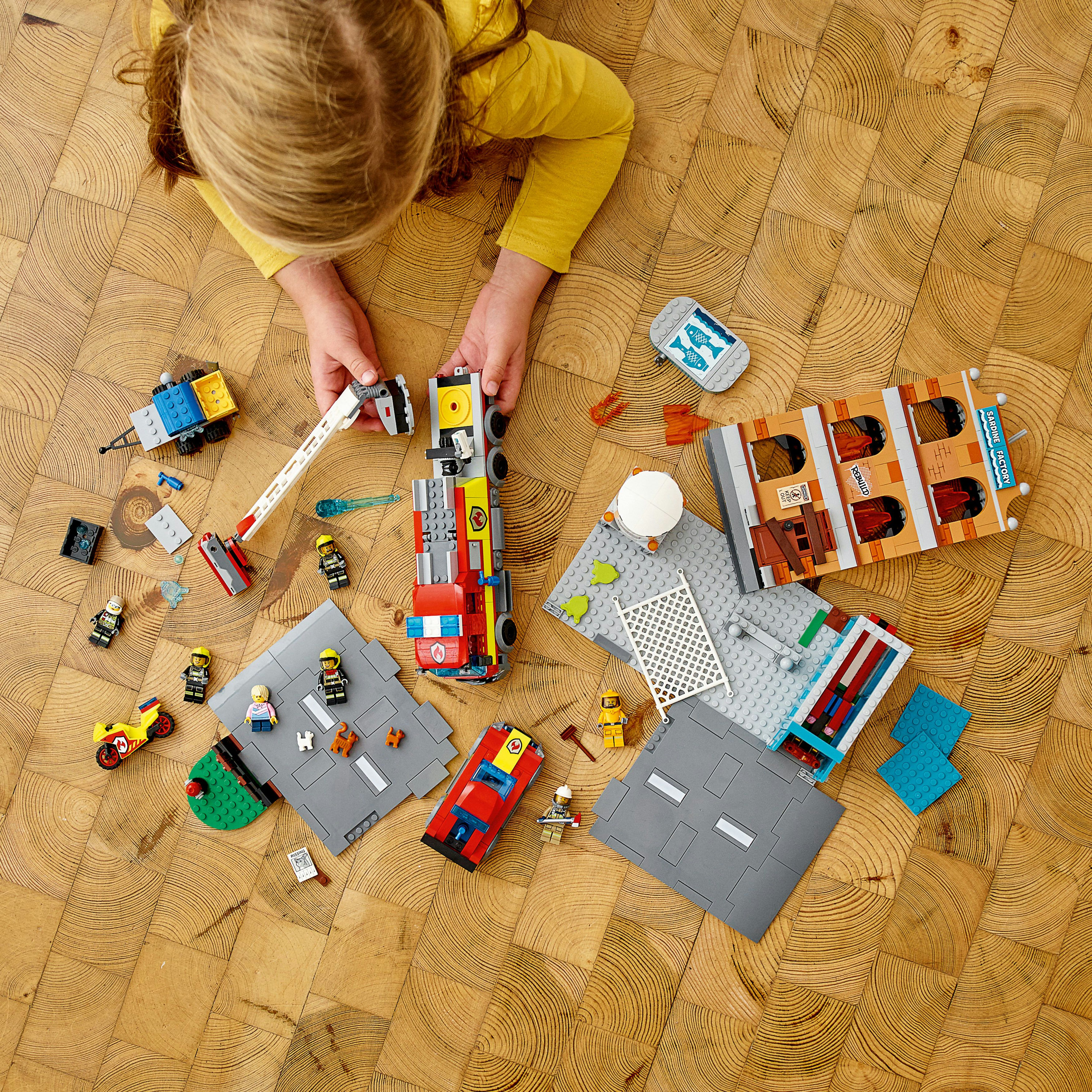 Un enfant jouant avec des pièces Lego, dont un camion de pompiers et un plan de bâtiment sur un sol en bois.