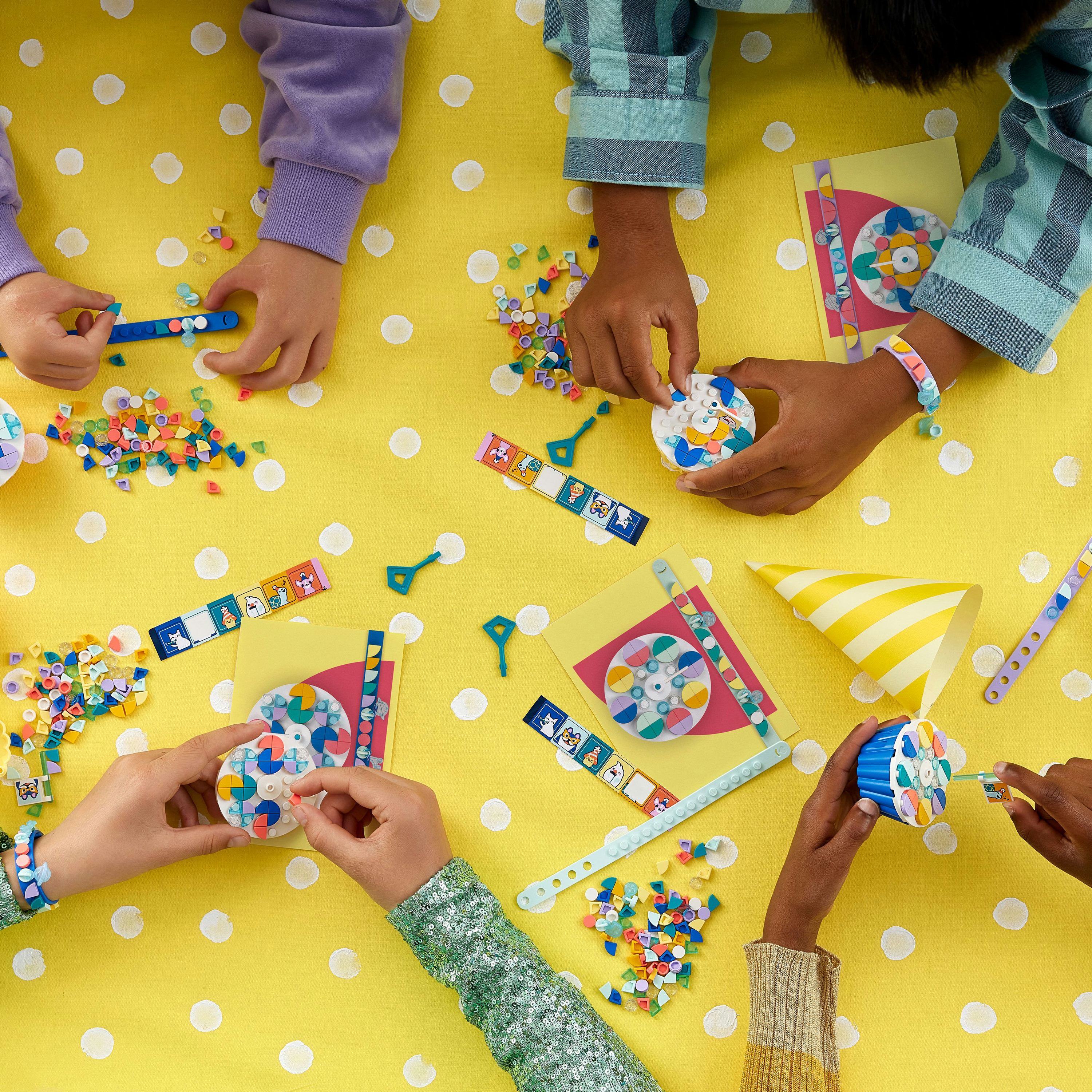 Hände, die bunte LEGO-Punkte auf einer gelben Oberfläche zusammensetzen. Perlen, Armbänder und Partyhüte sind auch zu sehen.