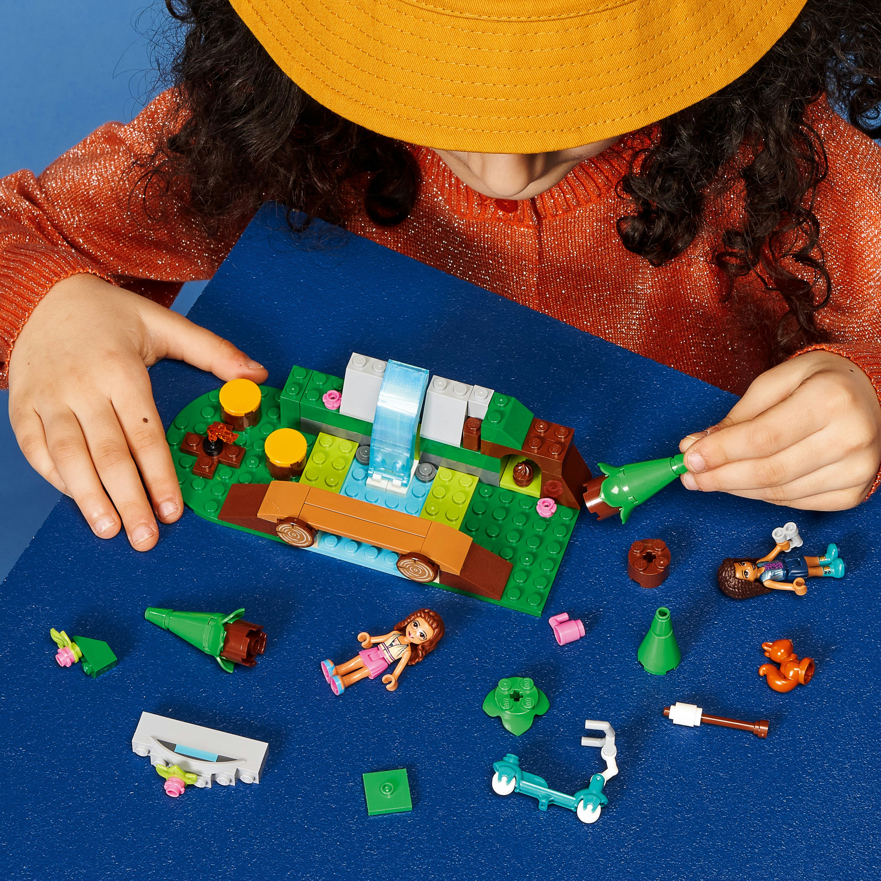 Ein Mädchen baut ein Lego-Set mit Wasserfall, Bäumen und Mini-Puppen auf blauem Hintergrund.
