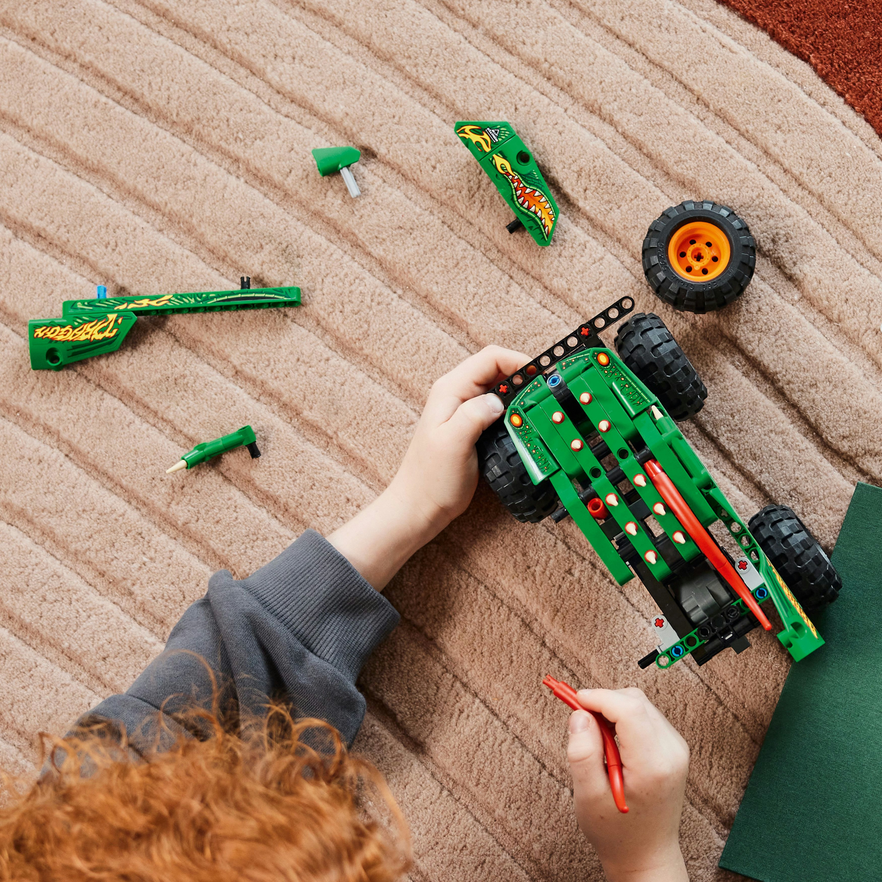 Mani di un bambino che assemblano un'auto Lego verde su un tappeto fantasia. Varie parti Lego sparse.