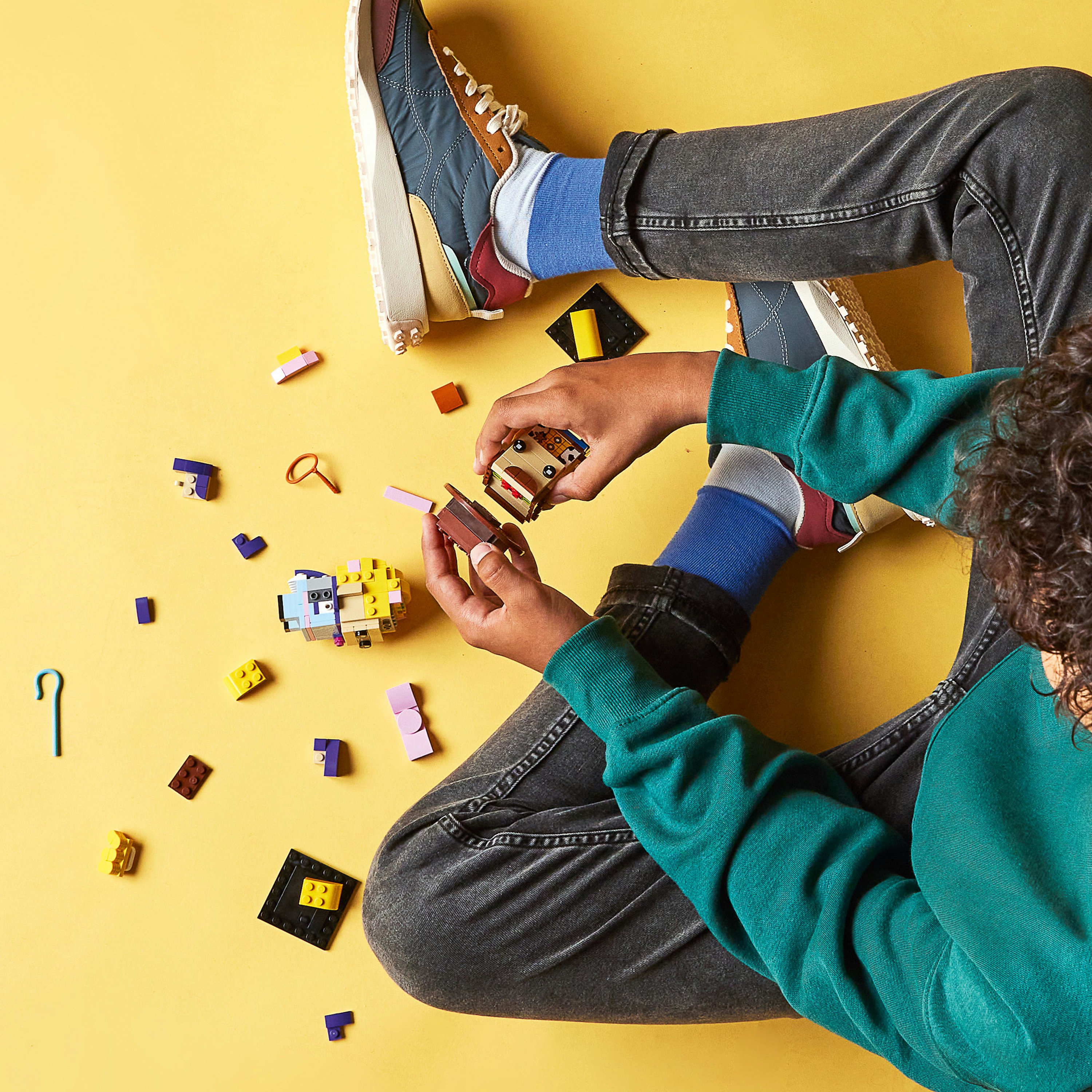 Eine Person mit verschränkten Beinen auf gelber Oberfläche, spielt mit verstreuten Lego-Steinen.