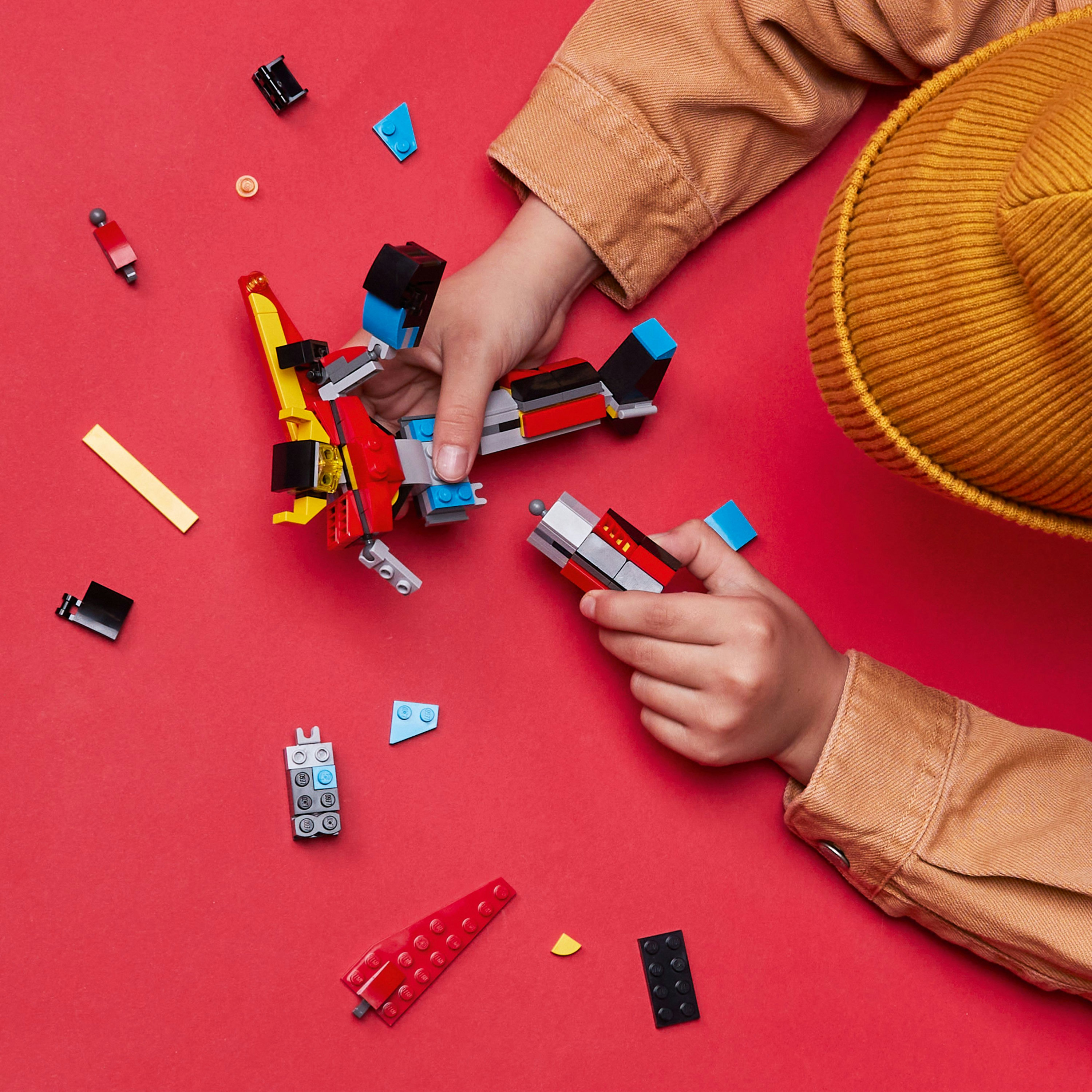 Manos ensamblando un avión Lego con piezas rojas y azules sobre fondo rojo.