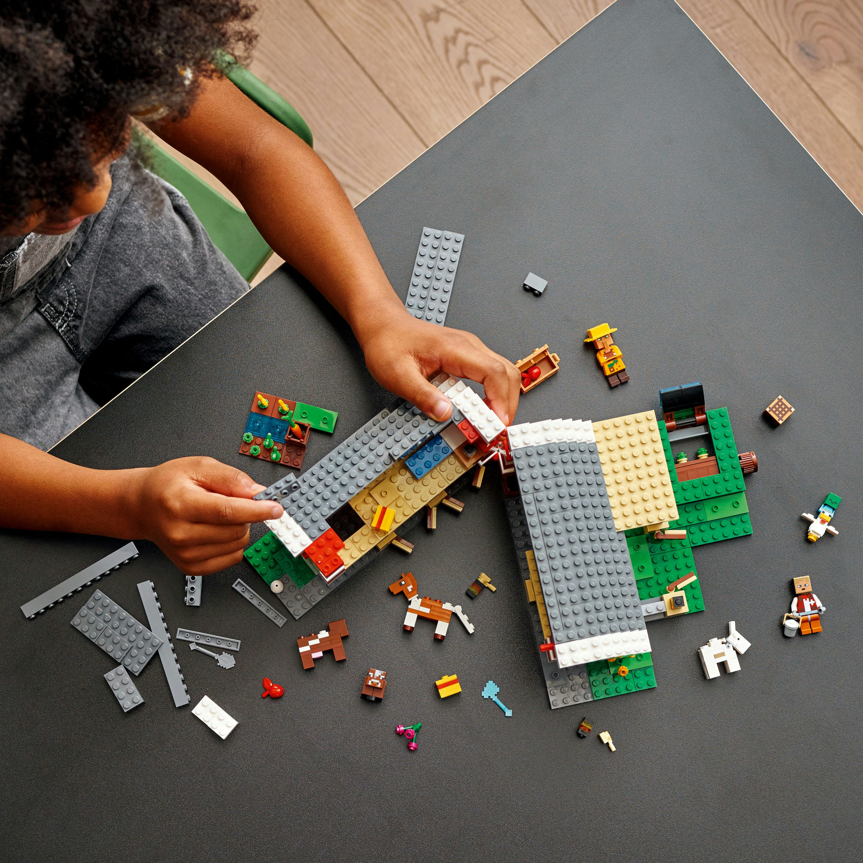 Kind baut mit Legosteinen. Grauer Tisch, Holzboden. Viele farbige Steine und Lego-Figuren sind vorhanden.
