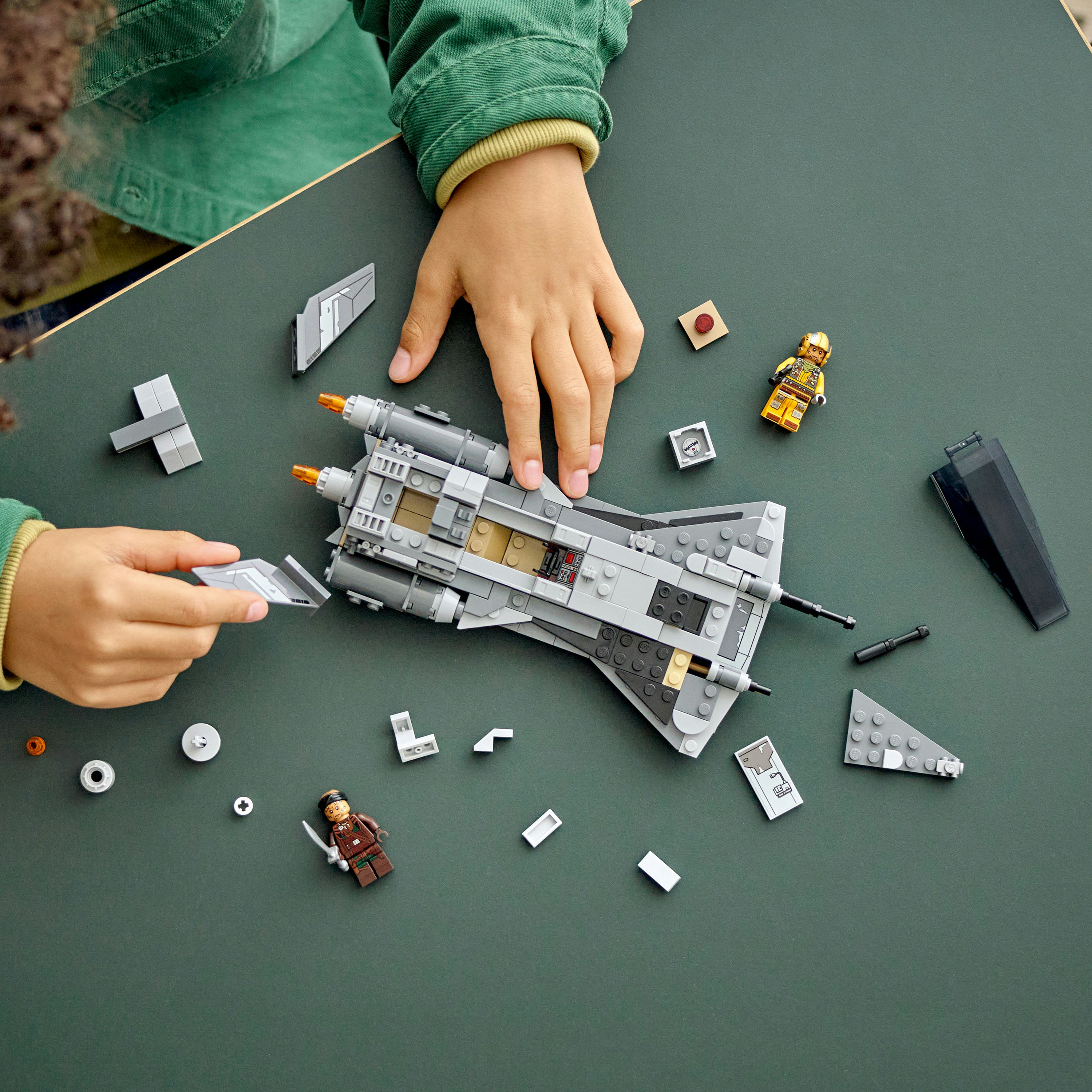 Manos ensamblan una nave espacial LEGO gris sobre una superficie verde. Figuras y piezas LEGO dispersas alrededor de la nave.
