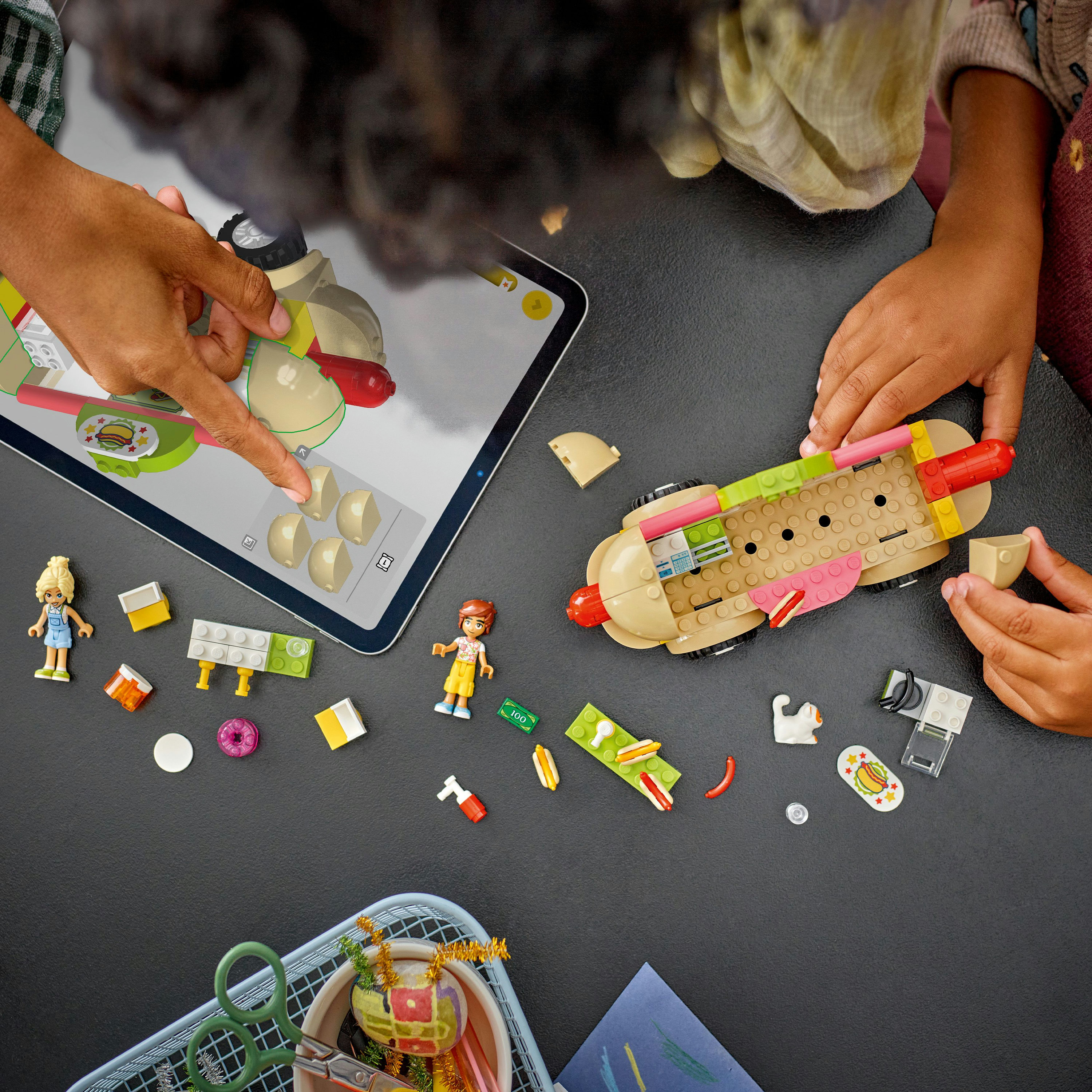 Niños construyendo un perrito caliente LEGO. Tableta muestra instrucciones. Piezas y minifiguras LEGO dispersas.