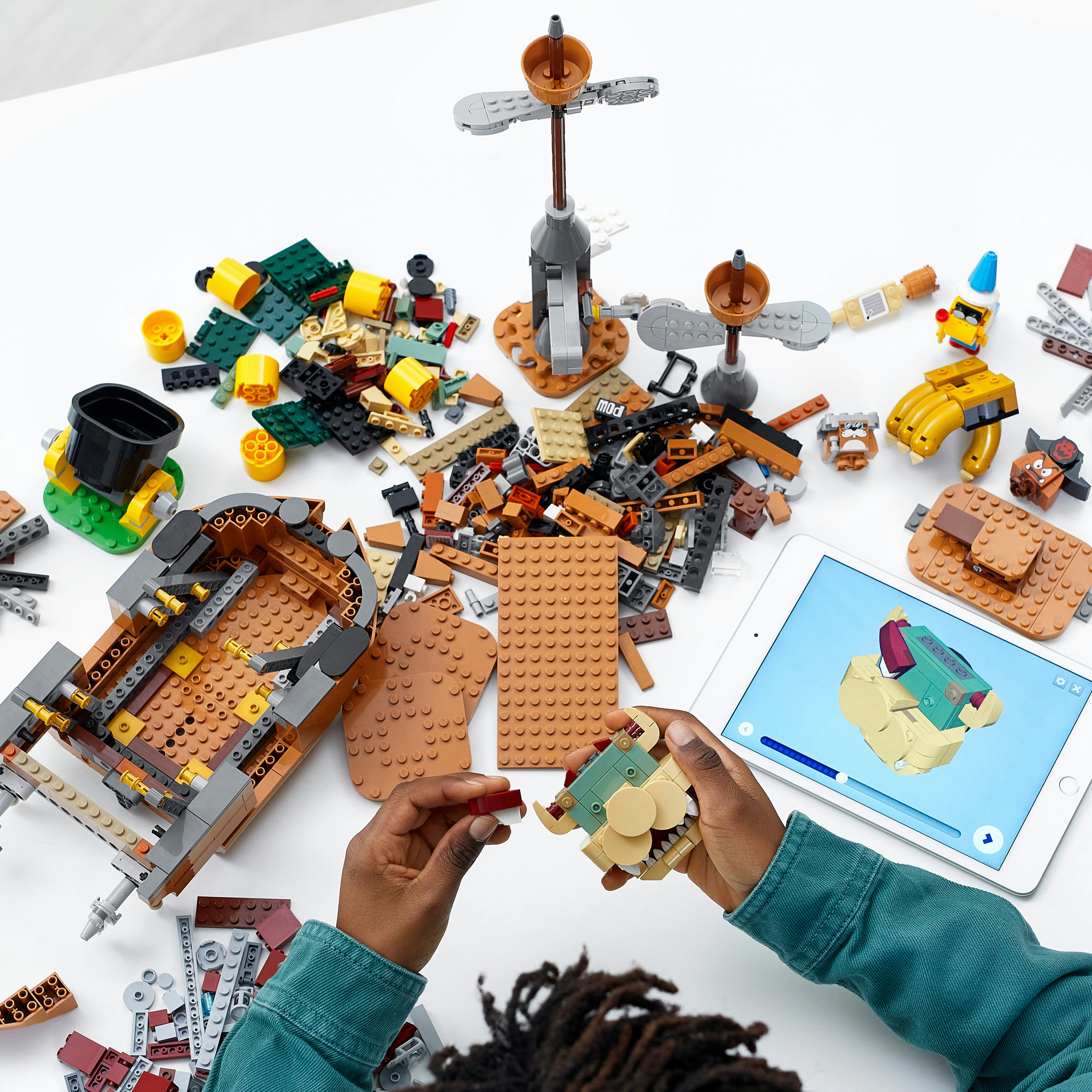 Mani che assemblano LEGO con un tablet. Vari pezzi LEGO sparsi su una superficie bianca.