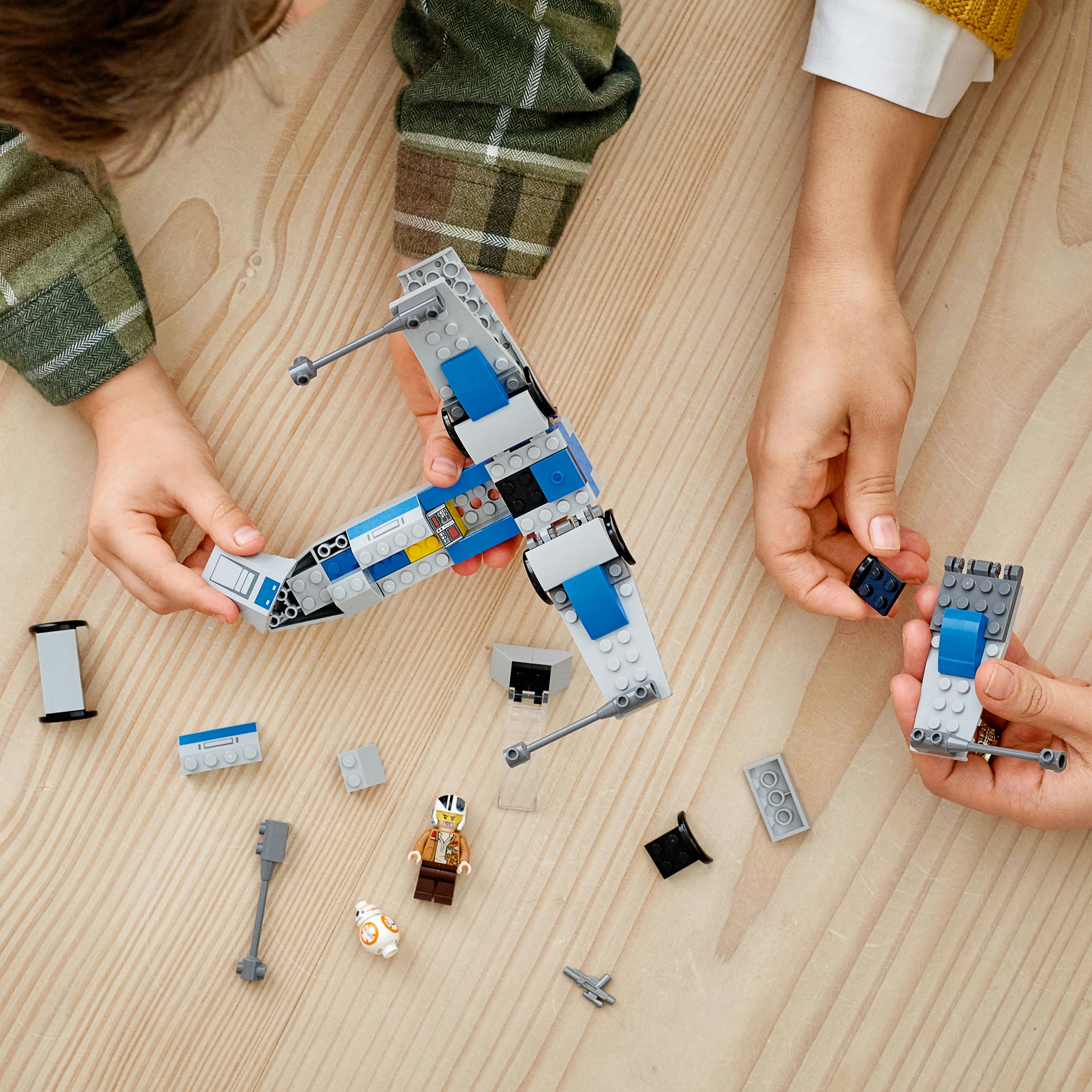Hände, die ein Lego X-Wing-Jäger-Modell zusammensetzen. Enthält Figuren und verstreute Lego-Teile auf Holzoberfläche.