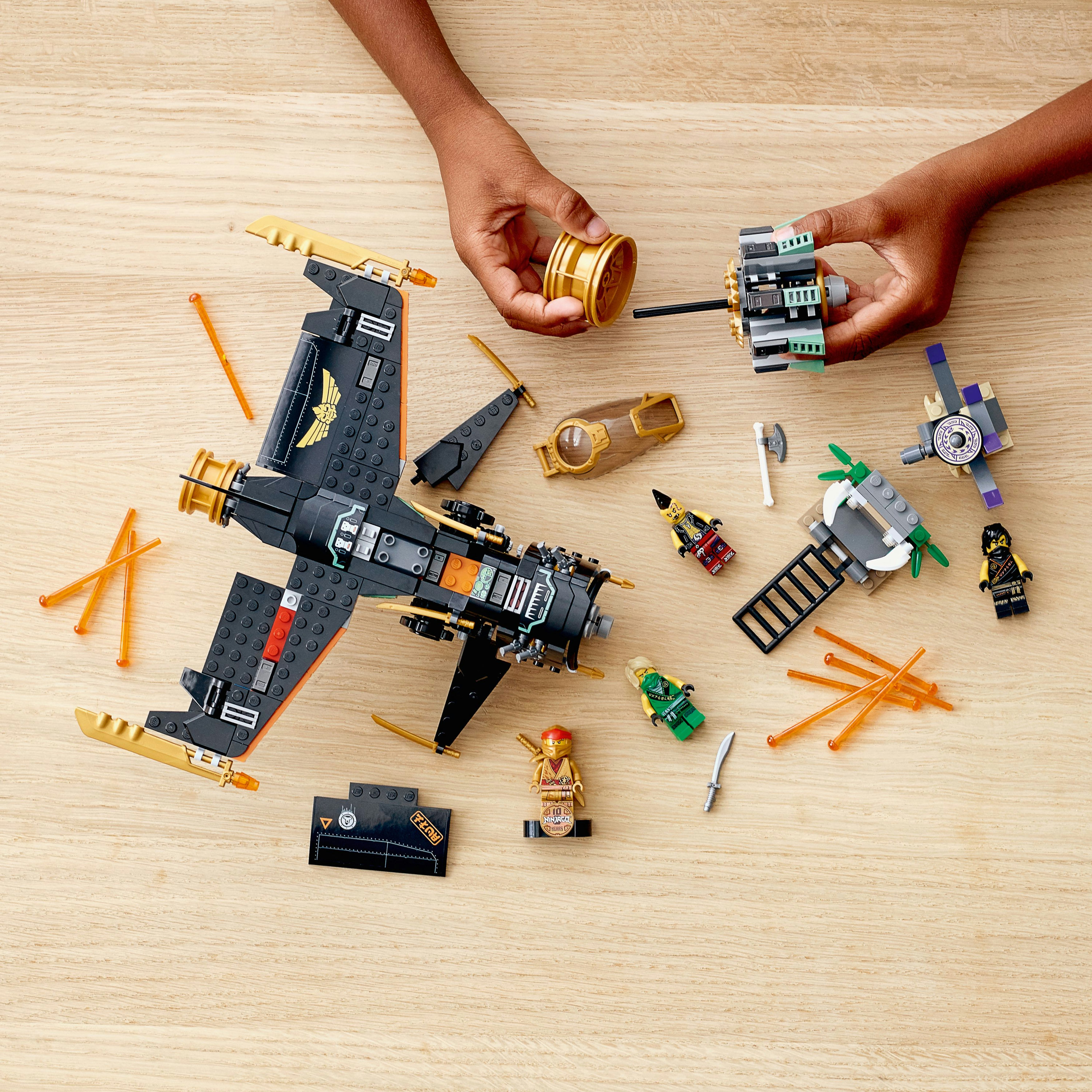 Hands are building a Lego model. The model includes an aircraft, minifigures, and various Lego pieces scattered around.