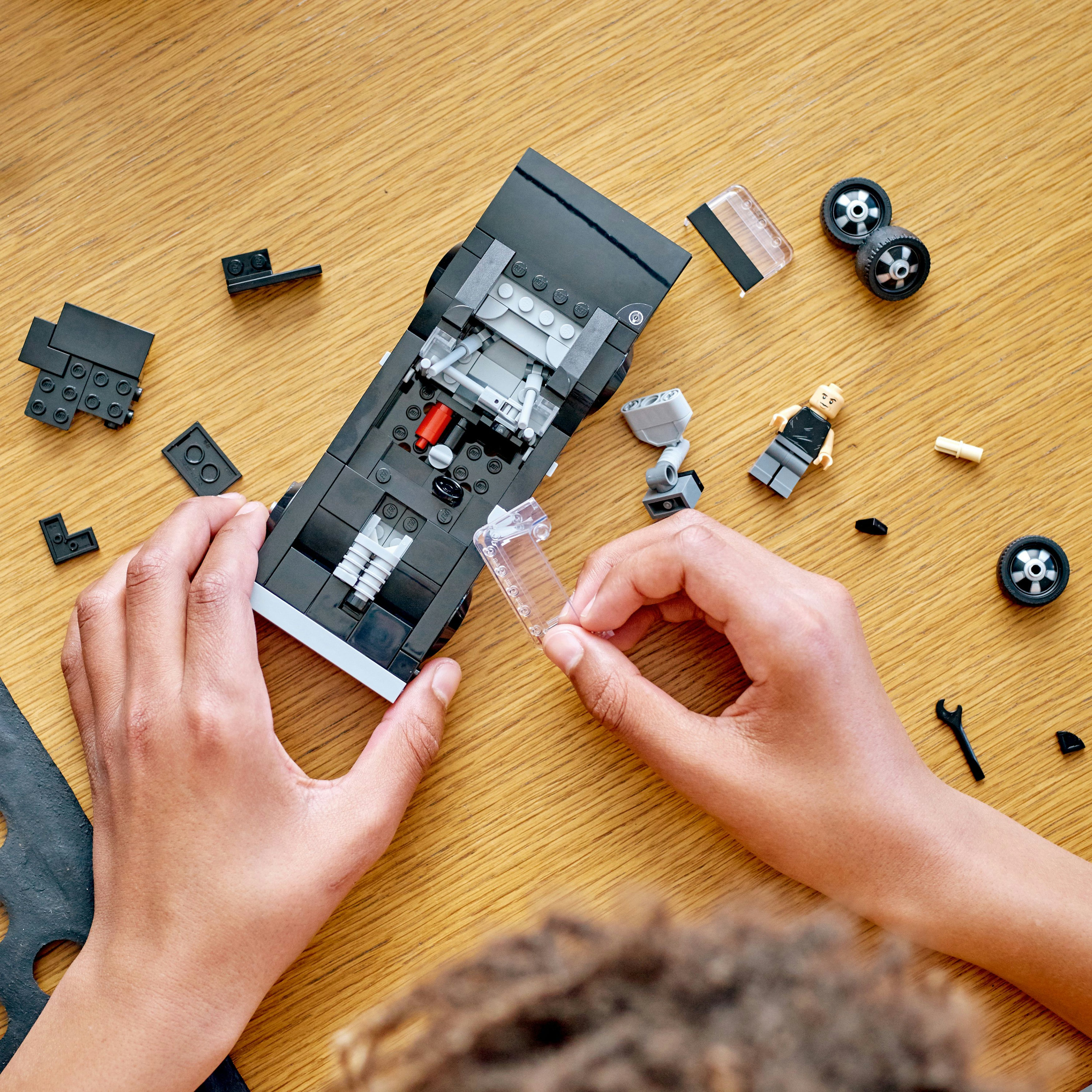 Ręce składające samochód Lego. Samochód jest czarny, z szarym wnętrzem i częściami na drewnianym stole.