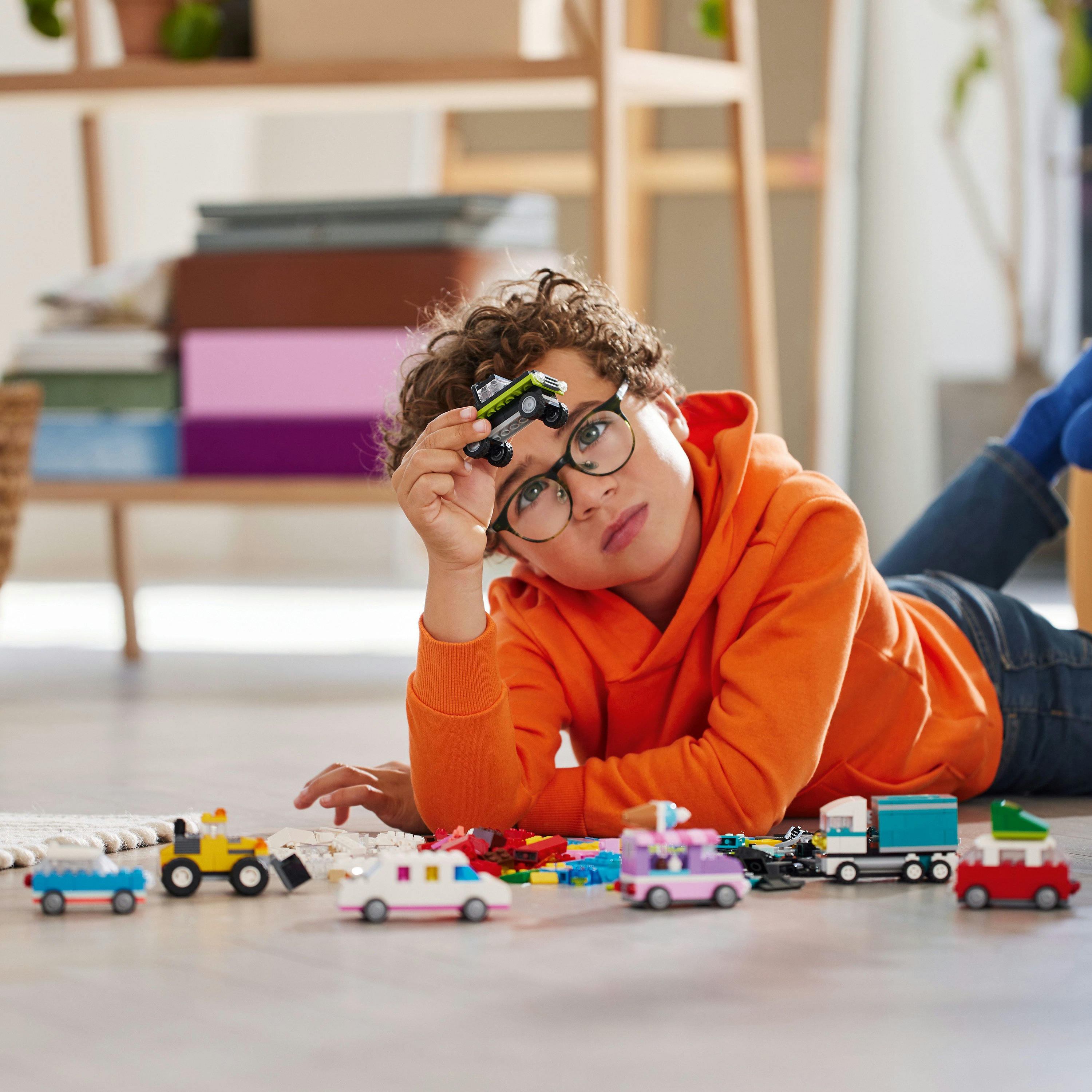 Un ragazzino sdraiato con macchinine Lego, tiene in mano una piccola auto verde e indossa gli occhiali.