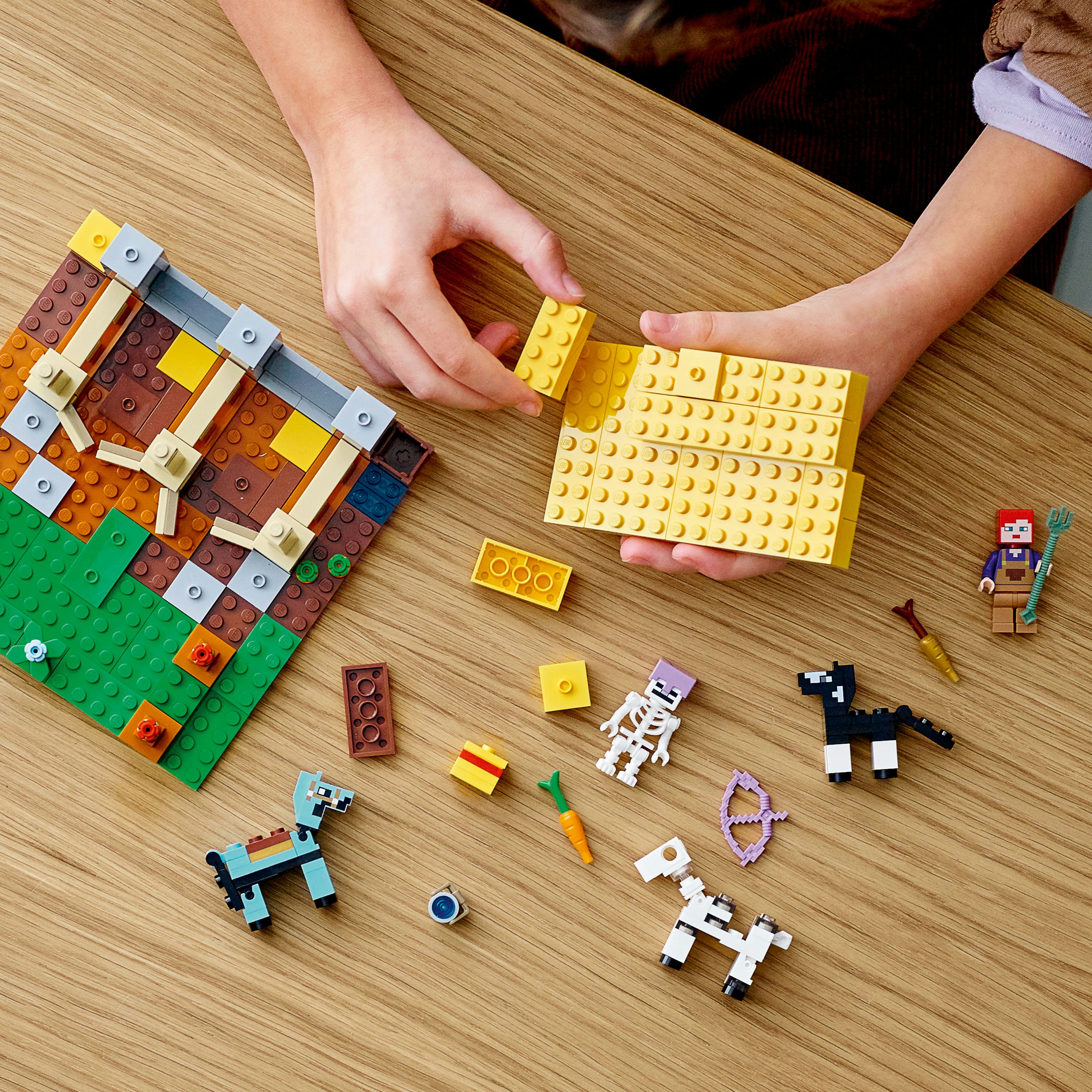 Mani che costruiscono una scena Lego Minecraft. Include blocchi, minifigure e piccole figure di animali, tutto su un tavolo di legno.