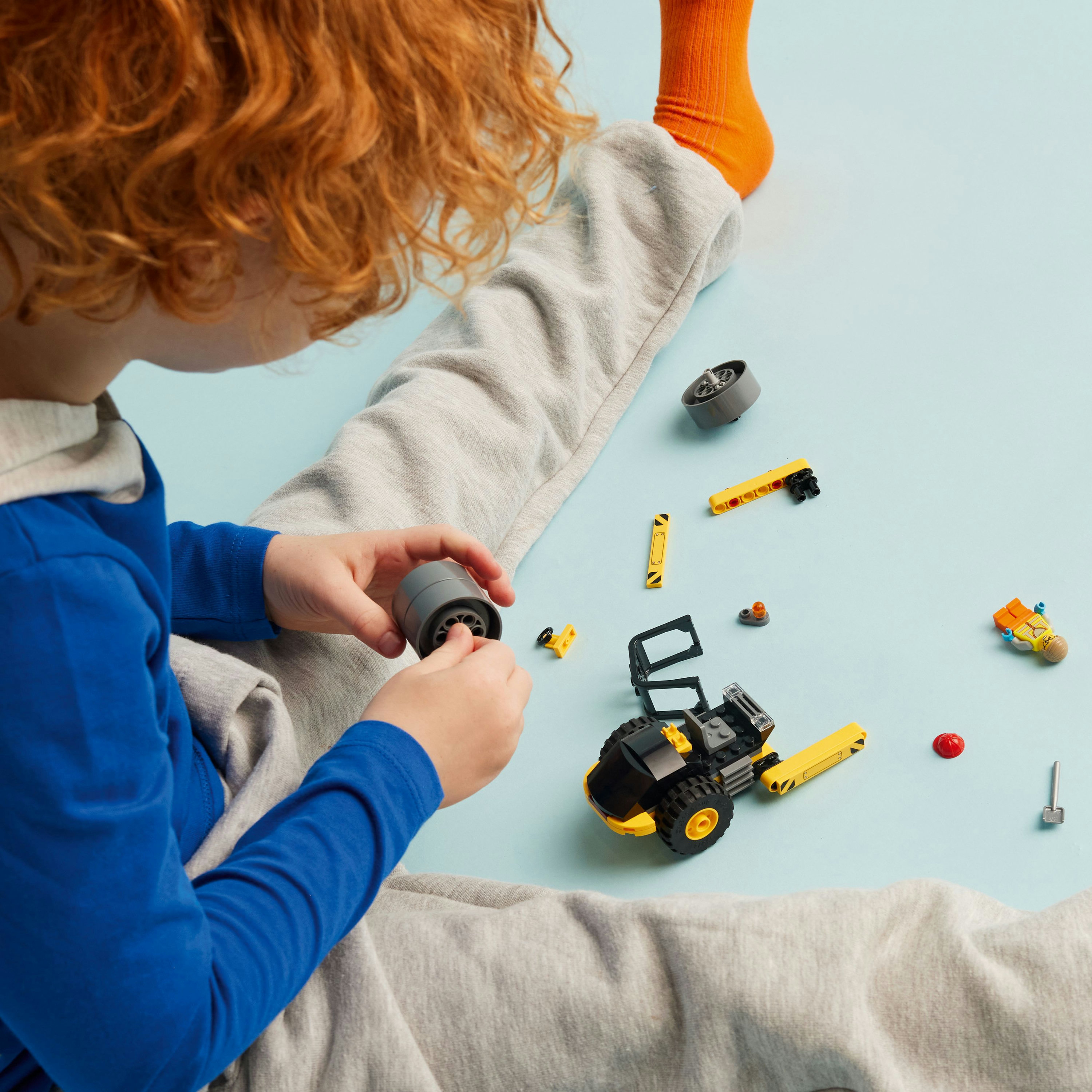 Een kind met een oranje sok speelt met Lego-stukken op een blauw oppervlak en monteert een klein voertuig.