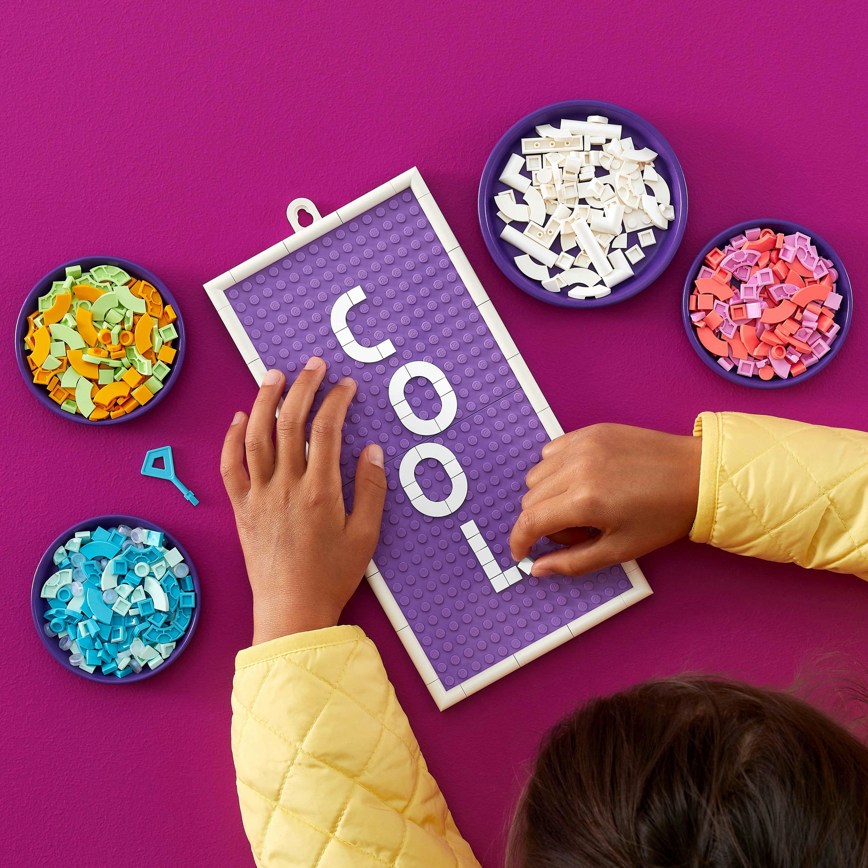Mani che dispongono mattoncini Lego su una base viola per scrivere "COOL", circondate da ciotole.