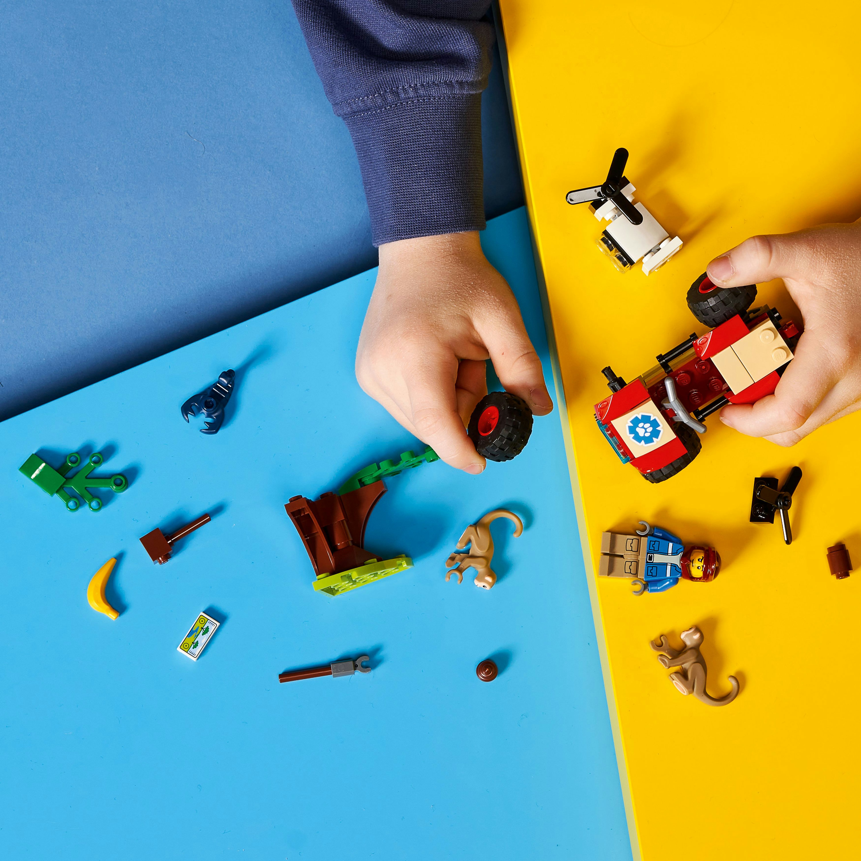 Kinderhände, die LEGO-Teile zusammensetzen. Ein rotes Auto, ein Affe, ein Baum und andere Spielelemente sind verstreut.