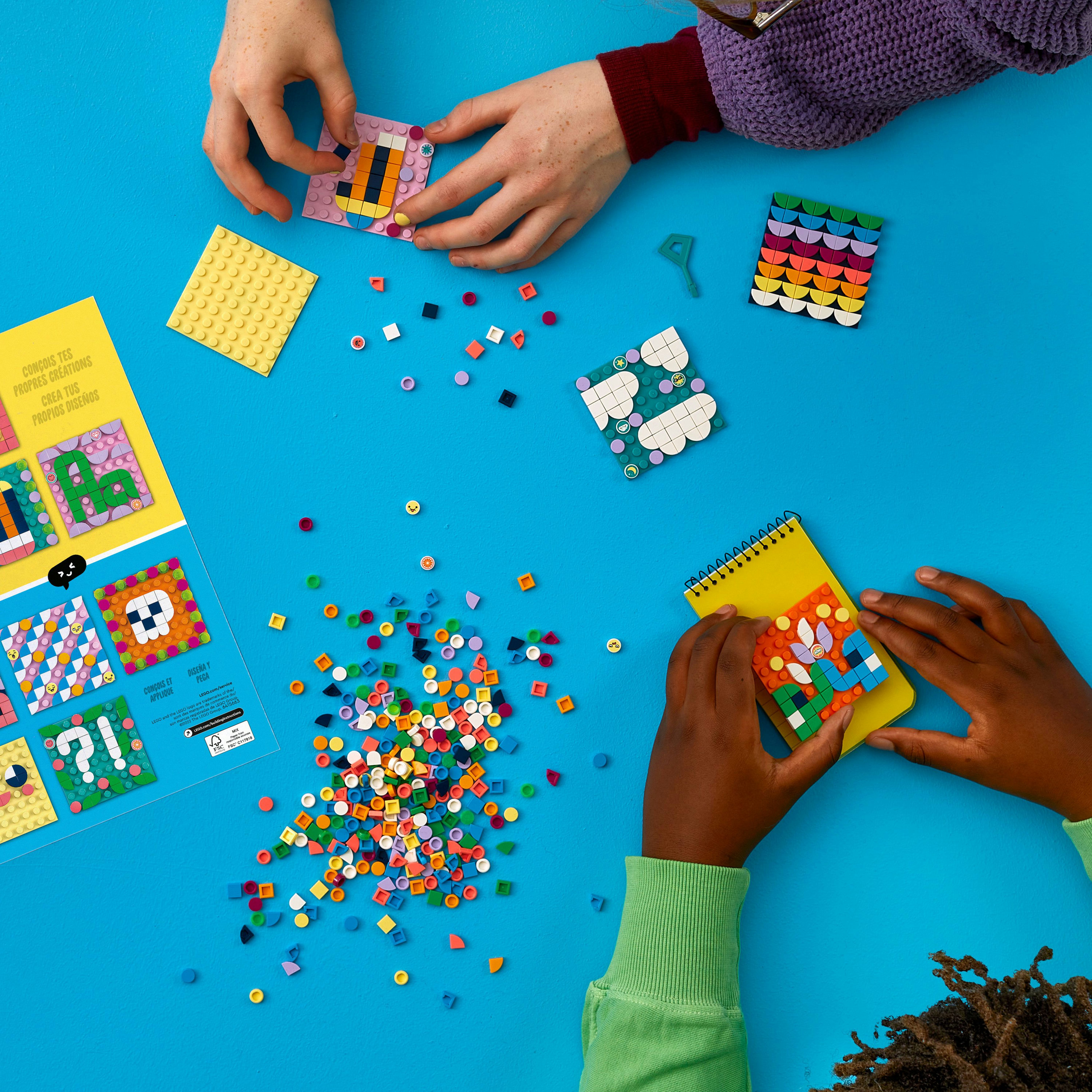 Hände bauen mit Lego Dots auf blauer Oberfläche, andere Dots und Anleitung.
