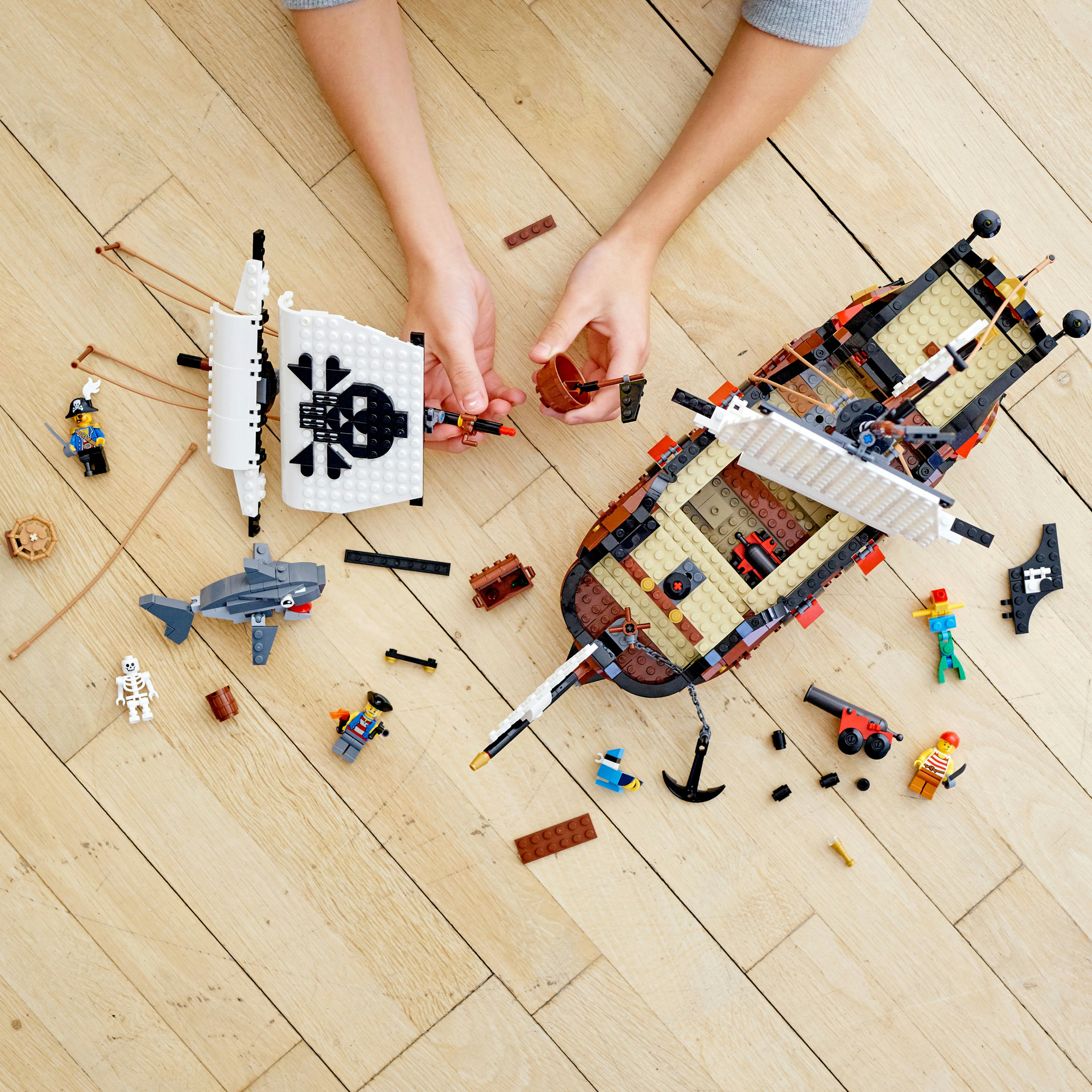 Ręce budujące statek piracki LEGO. Zawiera żagle, rekina, szkielet i minifigurki piratów na drewnianej podłodze.