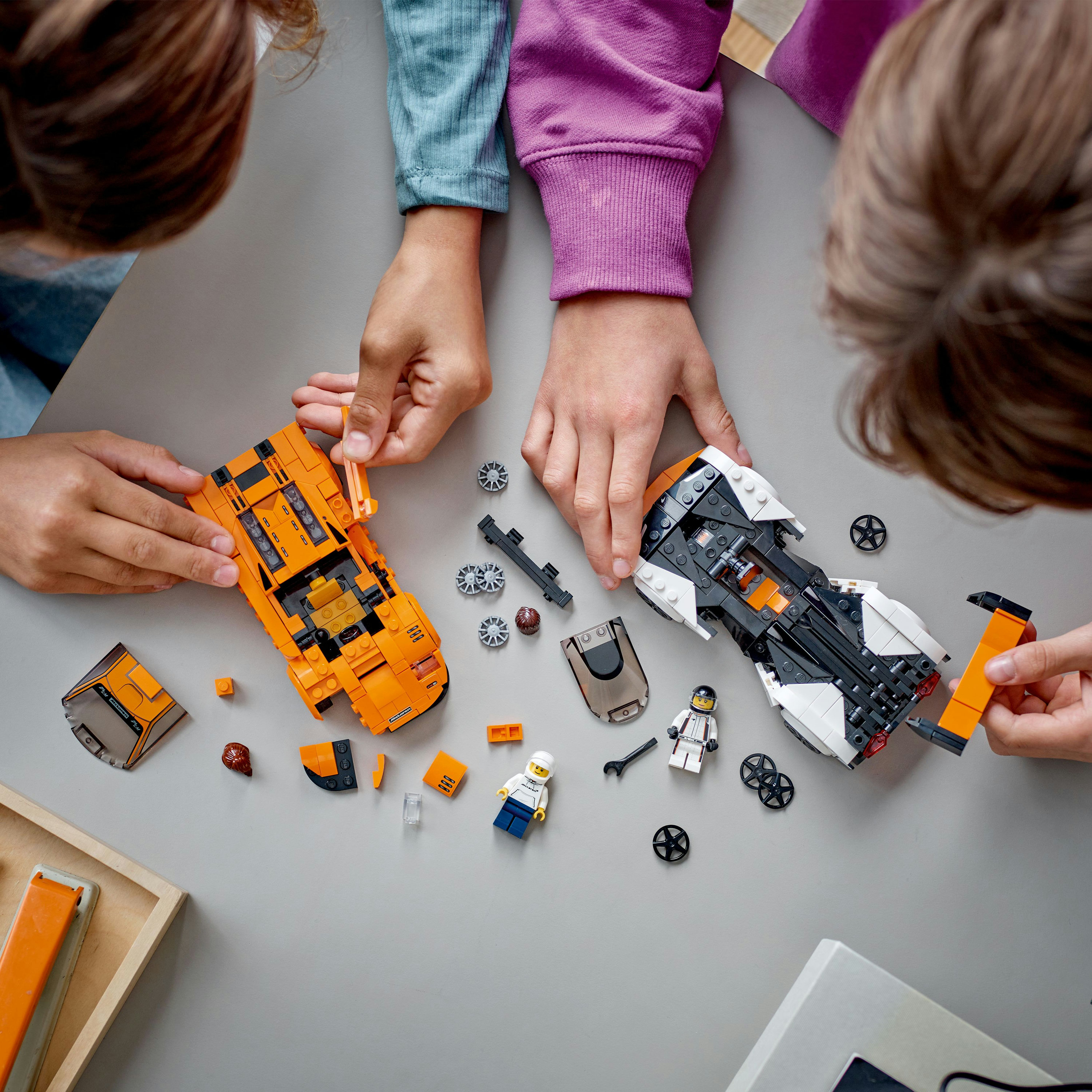 Zwei Kinder bauen orangefarbene und weiße Lego-Rennwagen auf einem grauen Tisch mit verstreuten Legoteilen.