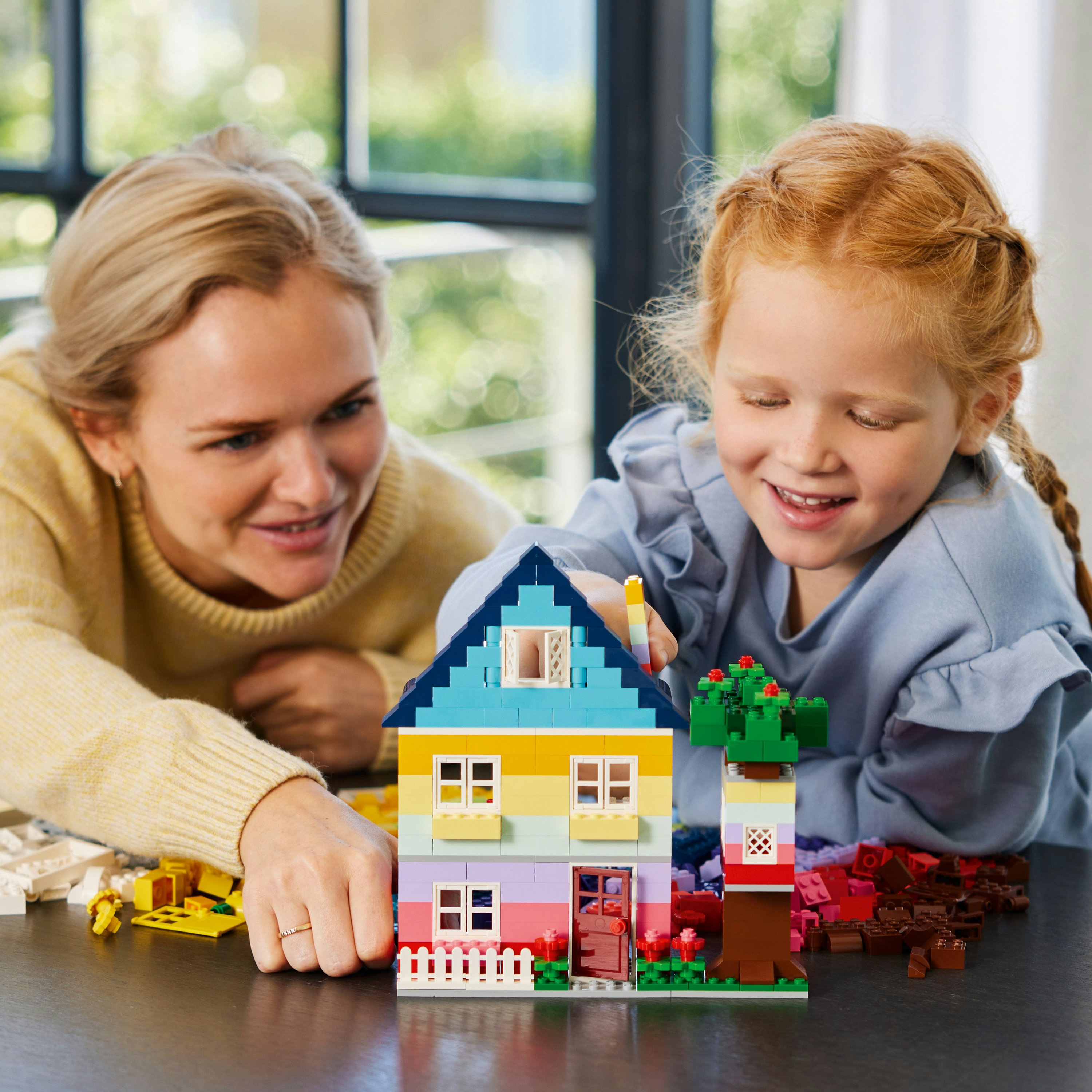 Una donna e una bambina costruiscono una casa Lego colorata. La casa ha un tetto blu e un albero.