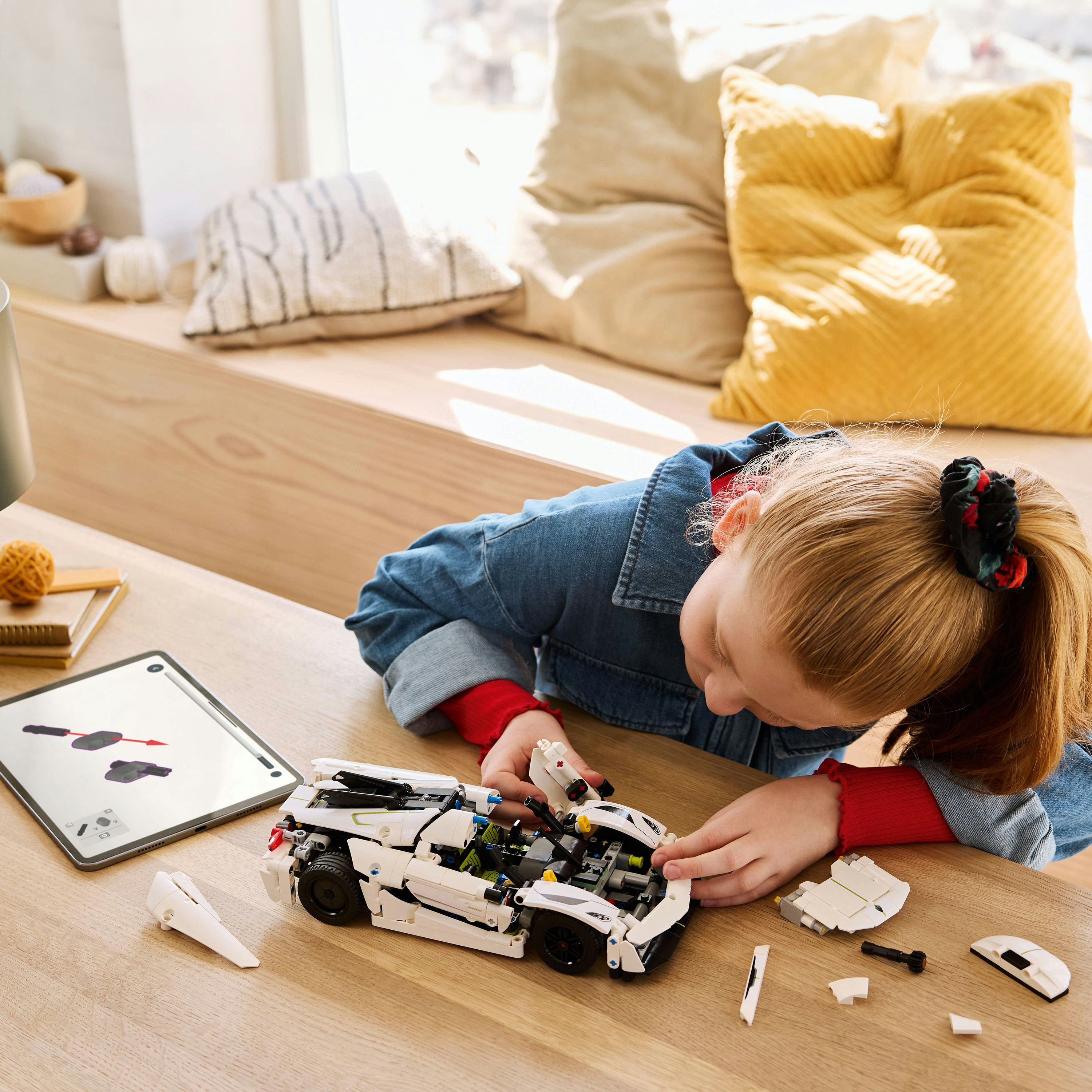 Une fille assemble un modèle de voiture Lego blanc sur une table en bois. Une tablette affiche des instructions.