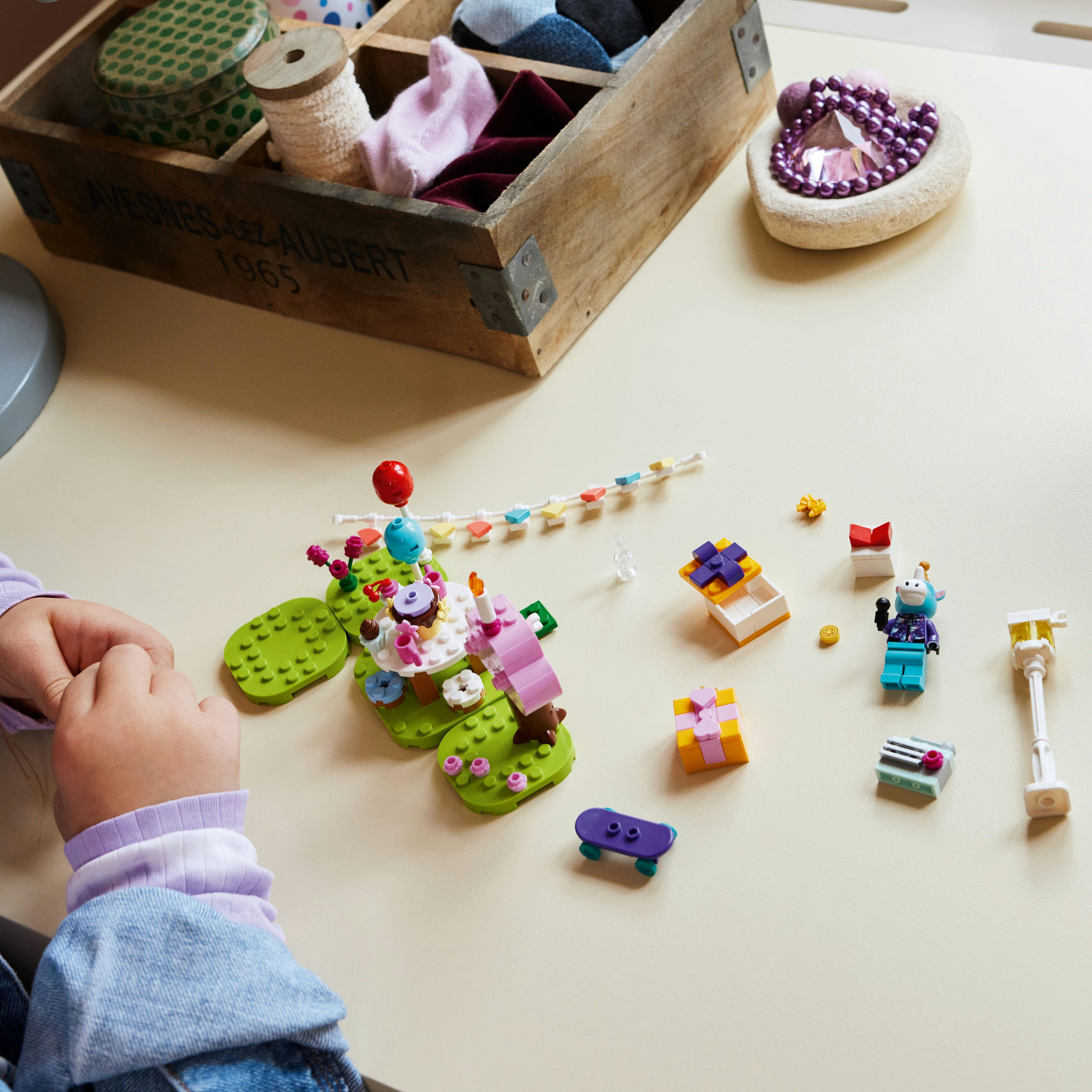 Çocuk, Lego parçalarıyla inşa ediyor. Doğum günü pastası ve hediye eşyaları. Küçük figürler ve mor bir kaykay mevcut.