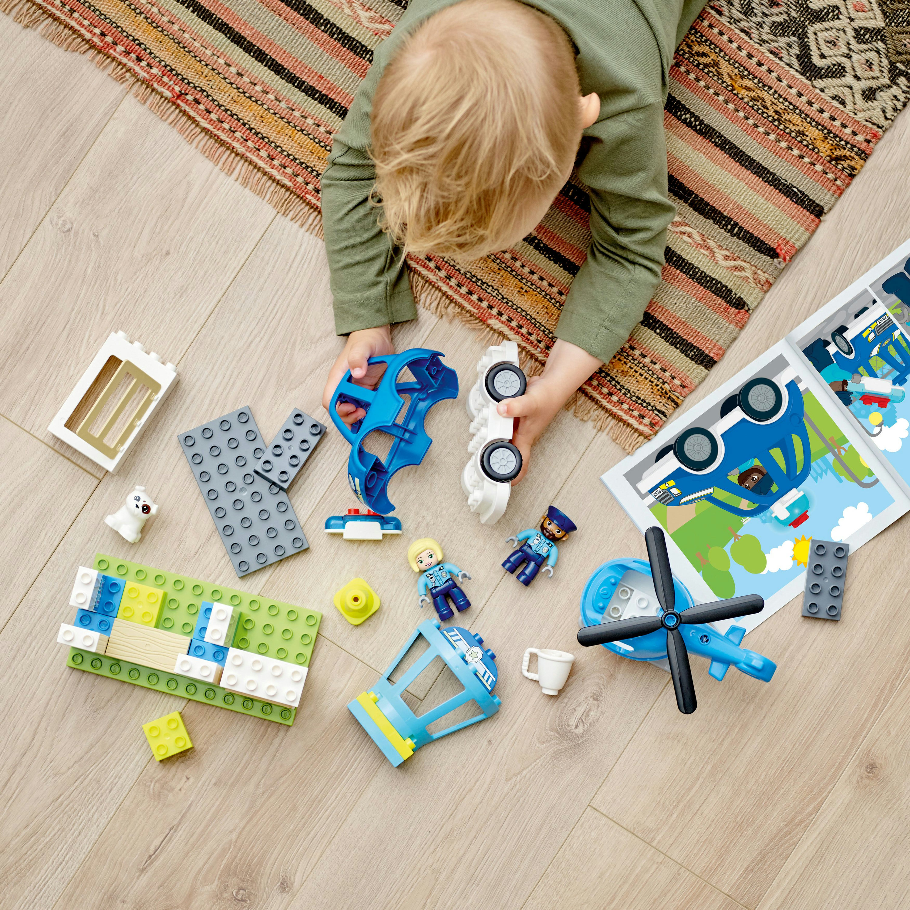 Niño jugando con bloques Duplo, piezas de coche y figuras. Alfombra y suelo de madera al fondo.