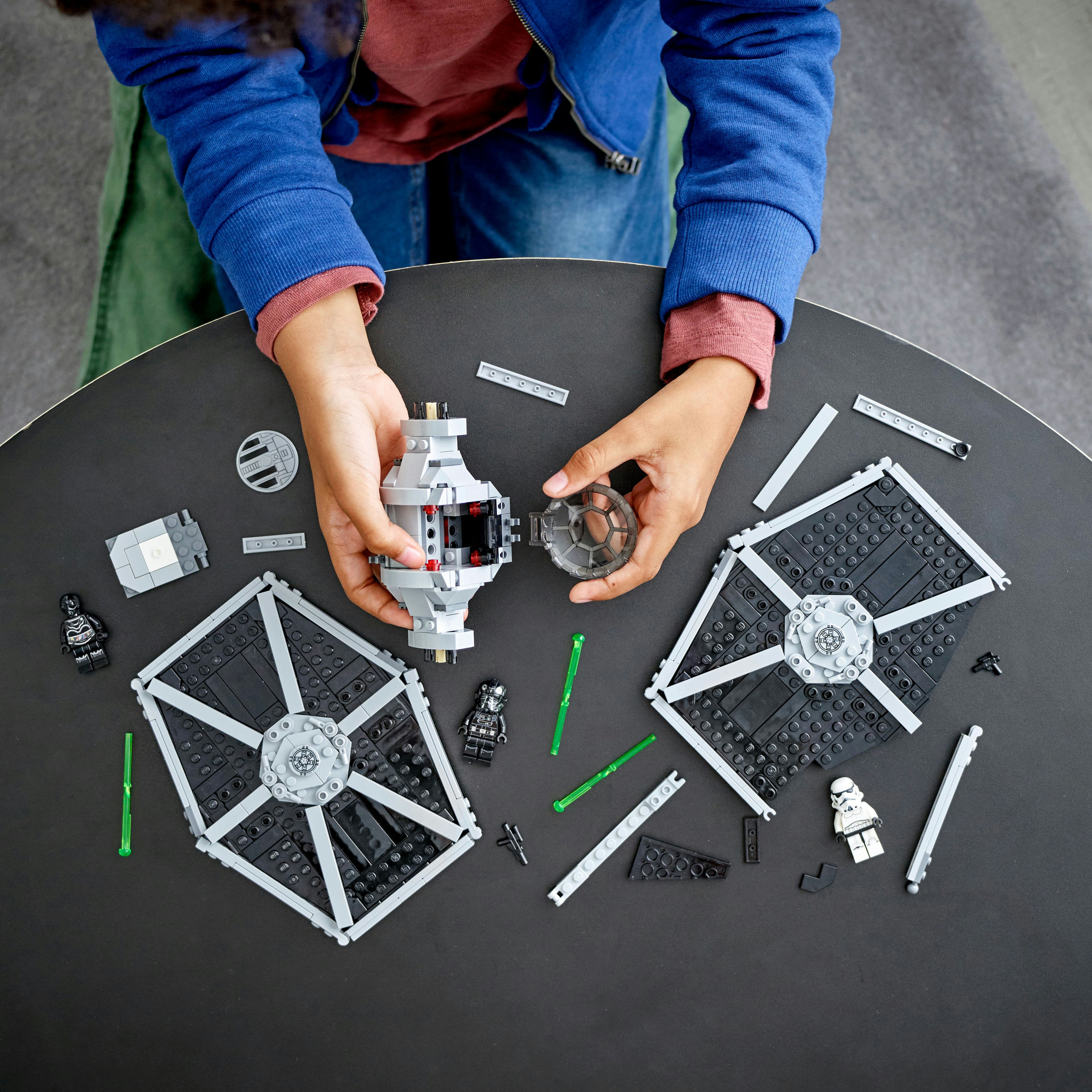 Bir çocuk, koyu bir yüzey üzerinde, dağınık parçalarla bir Star Wars Lego TIE fighter modeli topluyor.