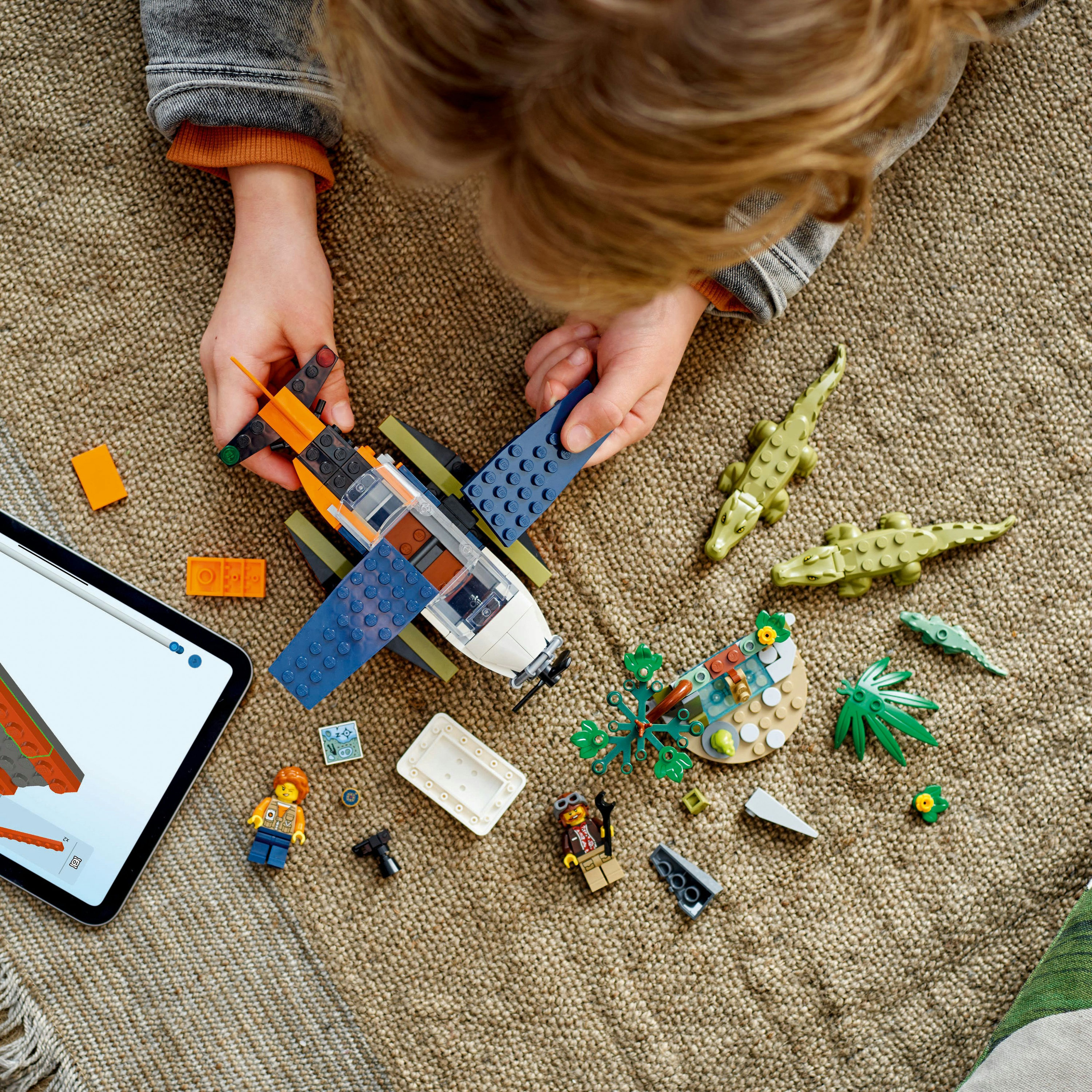 Bambino che gioca con un set Lego. Aereo, personaggi, coccodrilli, tablet, tappeto marrone. Vista dall'alto.