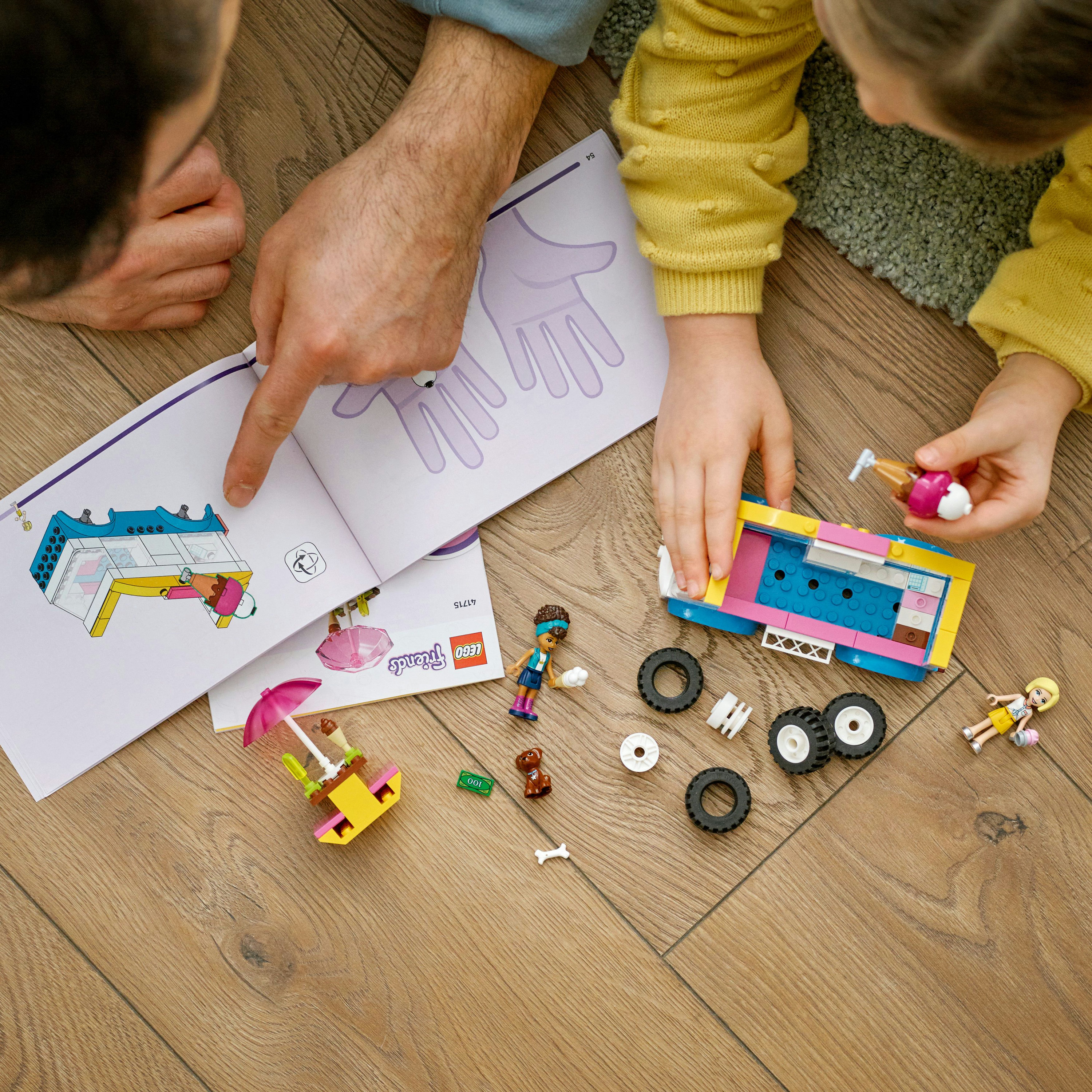 Hände zeigen auf eine Seite mit Lego-Anweisungen, zusammen mit Lego-Teilen und Figuren auf einer Holzoberfläche.