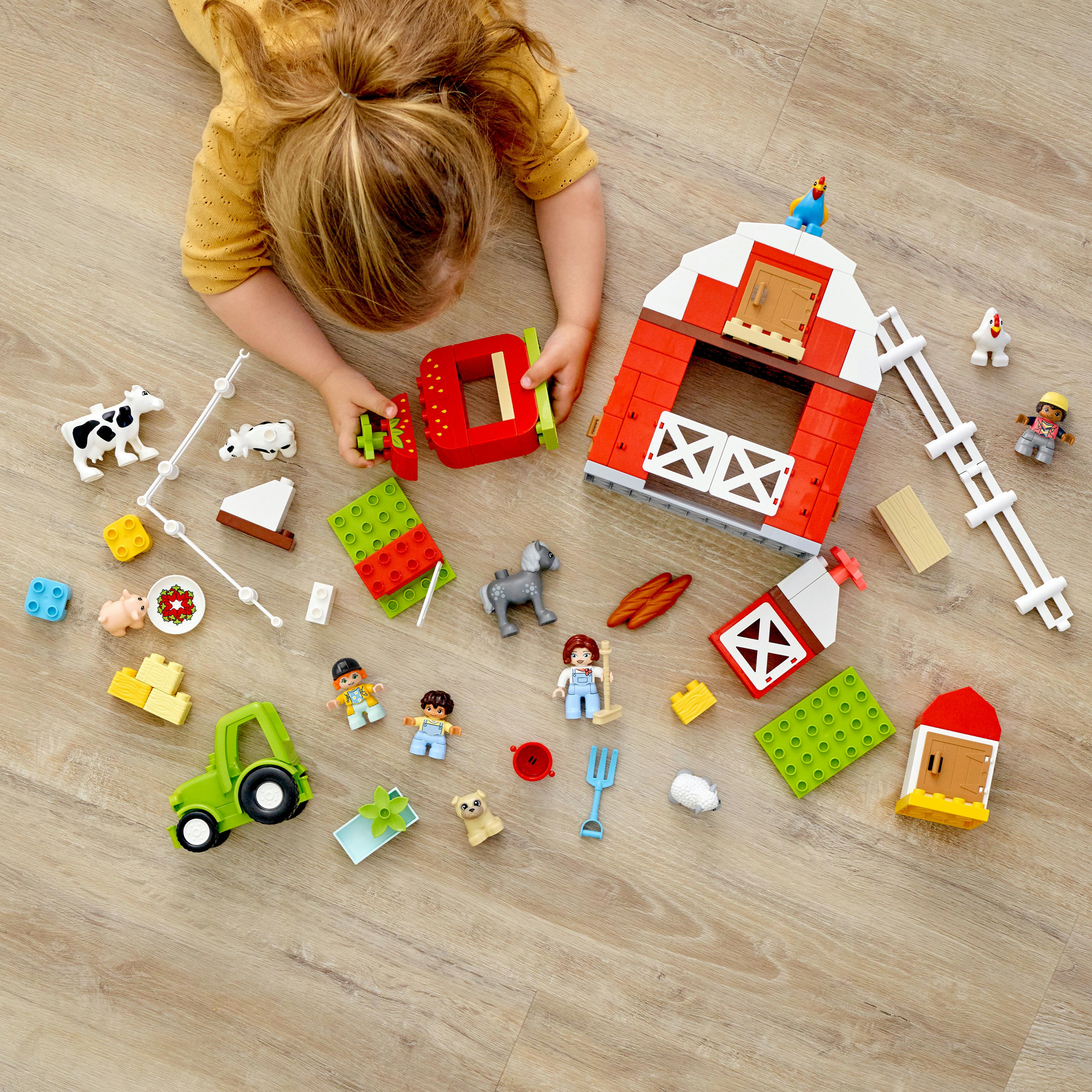 Ein Kind spielt mit einem Lego-Bauernhof-Set. Rote Scheune, grüner Traktor, Tiere und Blöcke verstreut auf Holzboden.