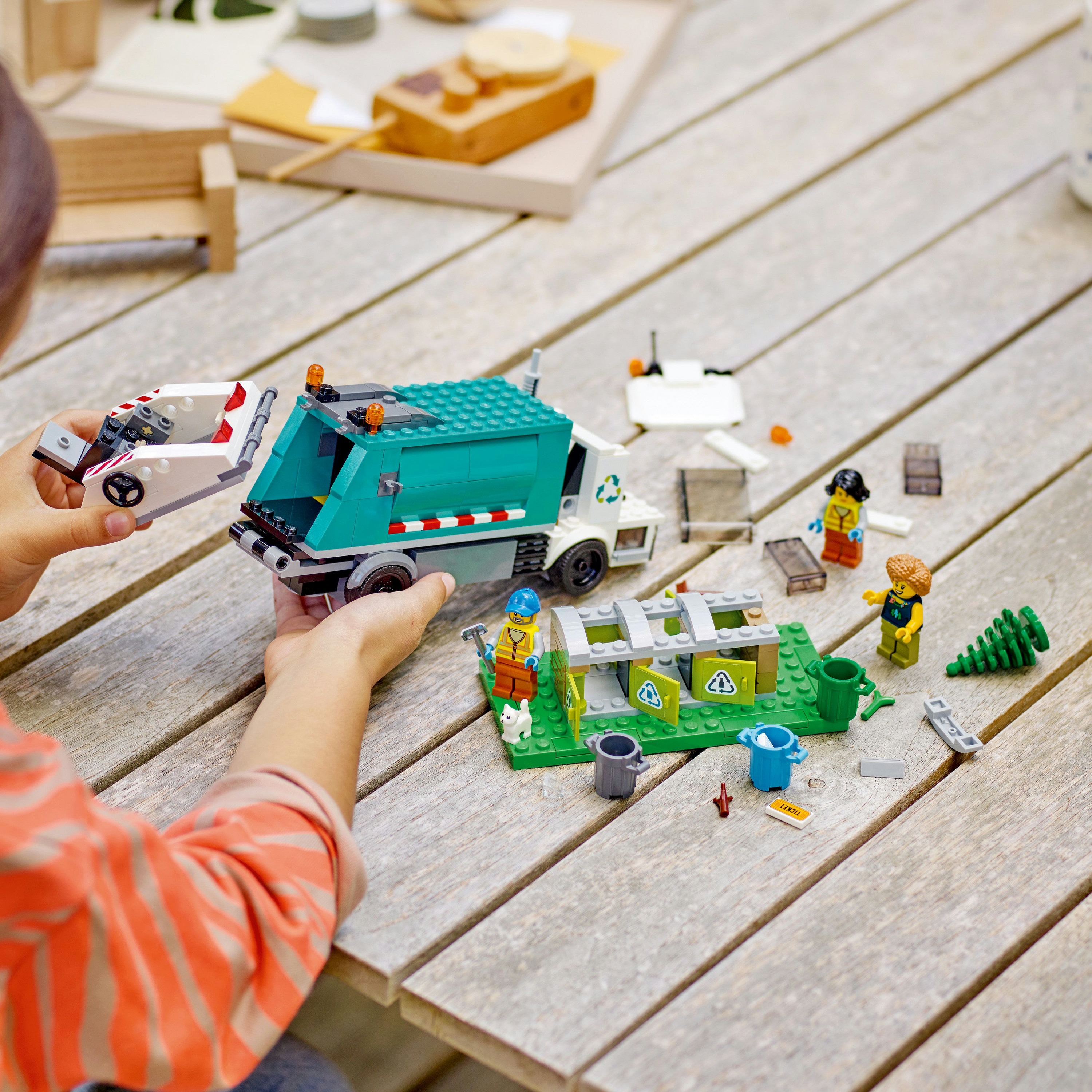 Kind spielt mit Spielzeug-Müllwagen und Lego-Figuren auf einem Holztisch im Freien.