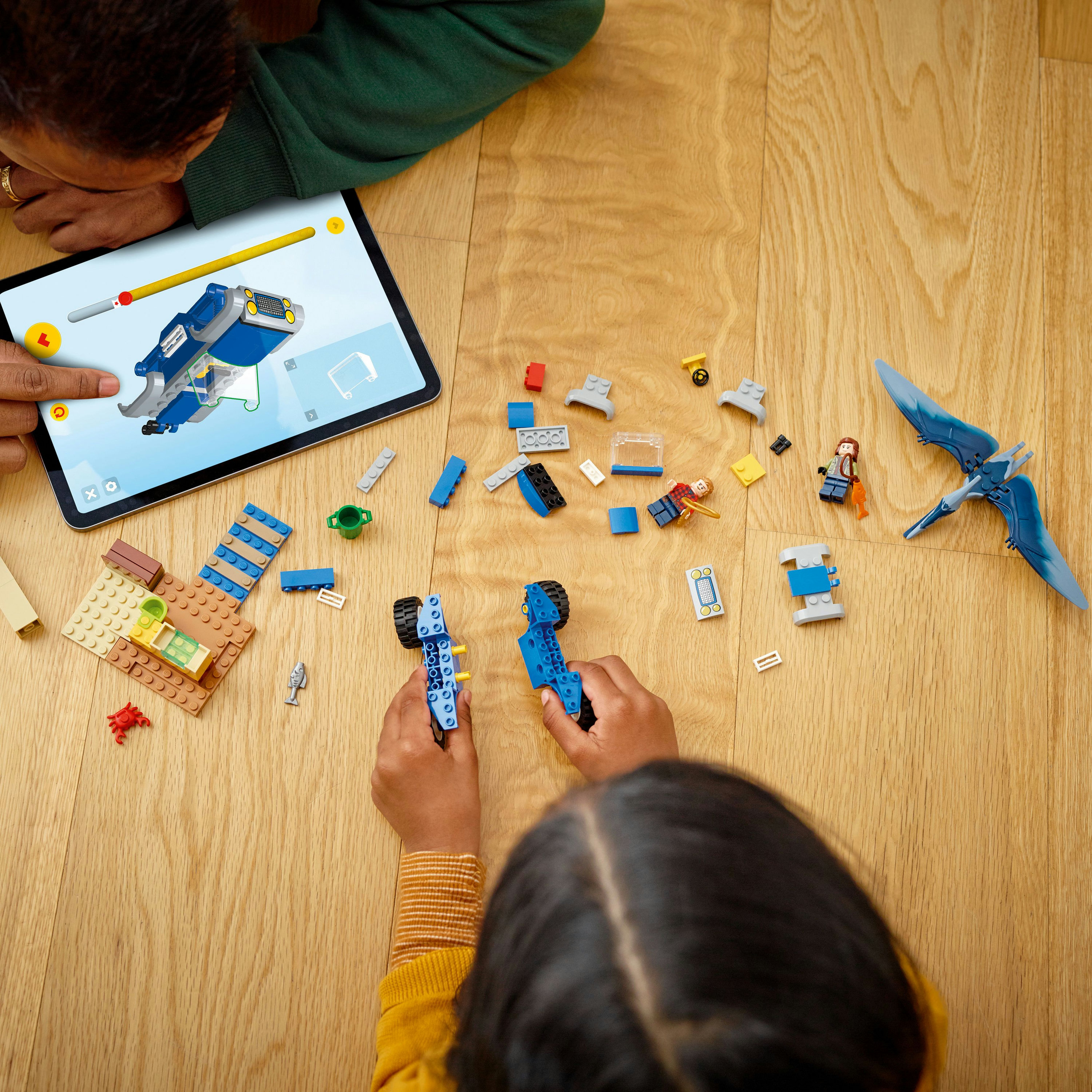 Bir çocuk mavi Lego araç yapıyor. Tablet talimatları gösteriyor. Diğer Lego parçaları dağınık.