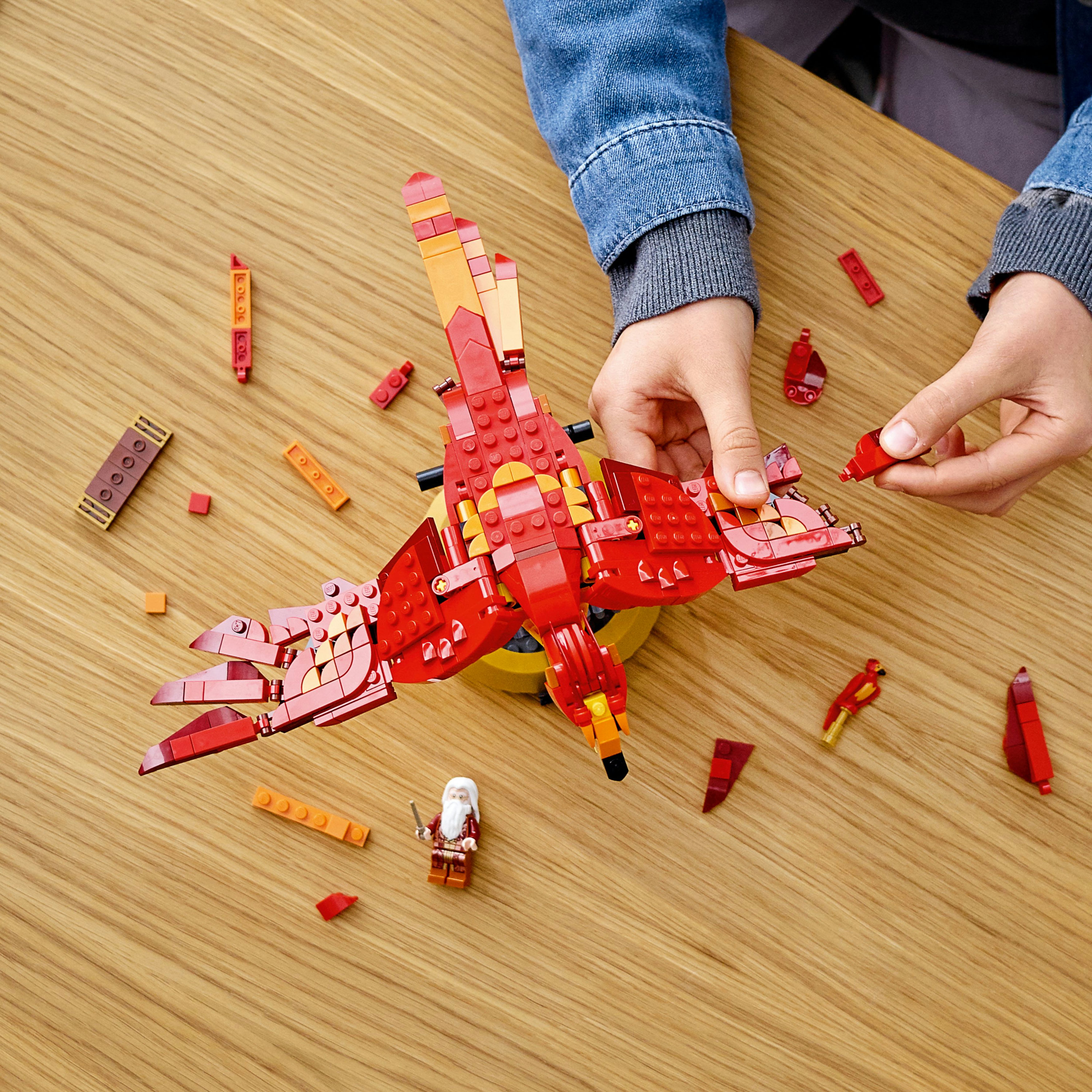 Mani che costruiscono una fenice Lego rossa, con pezzi sparsi su un tavolo di legno, vicino a una minifigure.