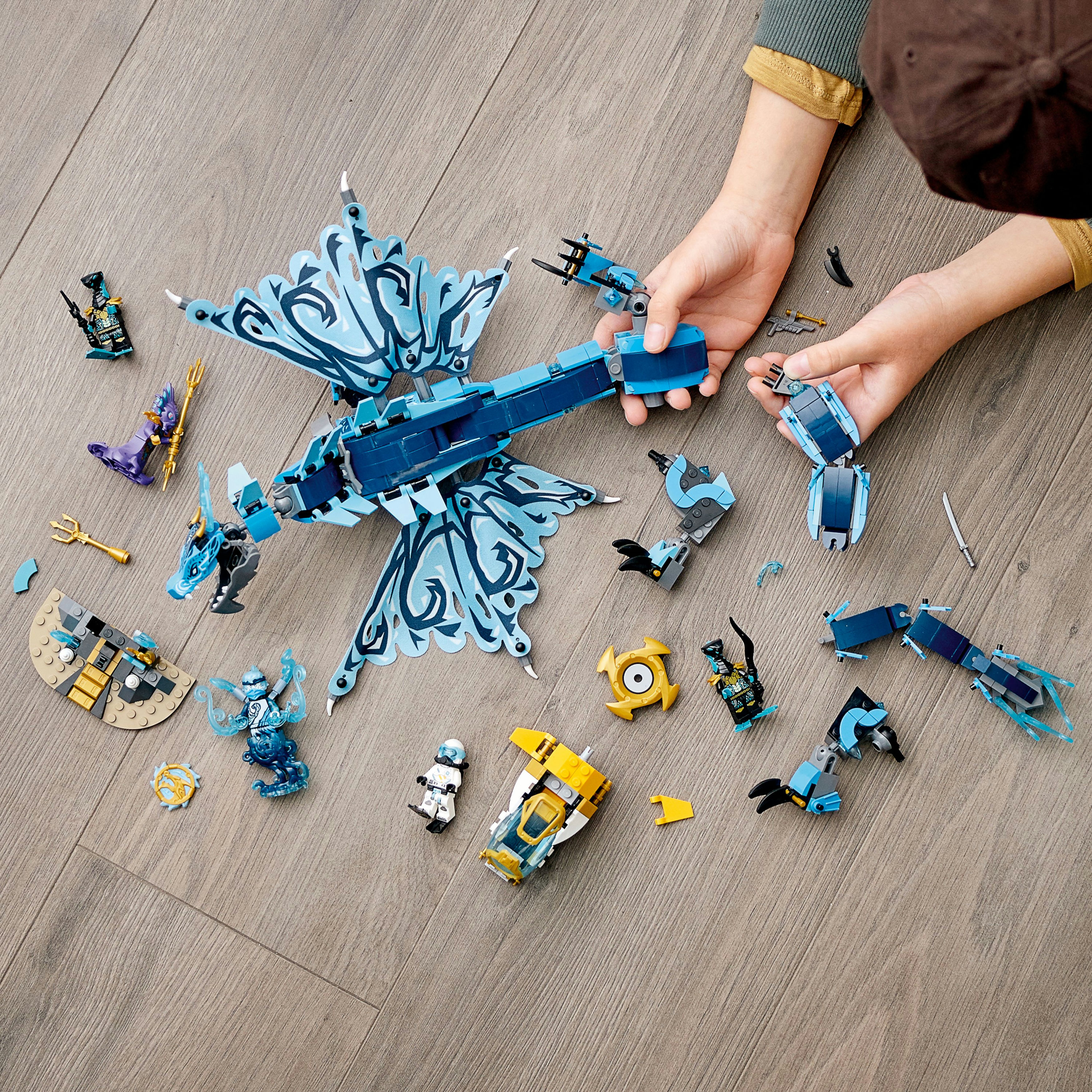 Mani che assemblano il drago Lego Ninjago, con minifigure e pezzi aggiuntivi sparsi sul pavimento di legno.