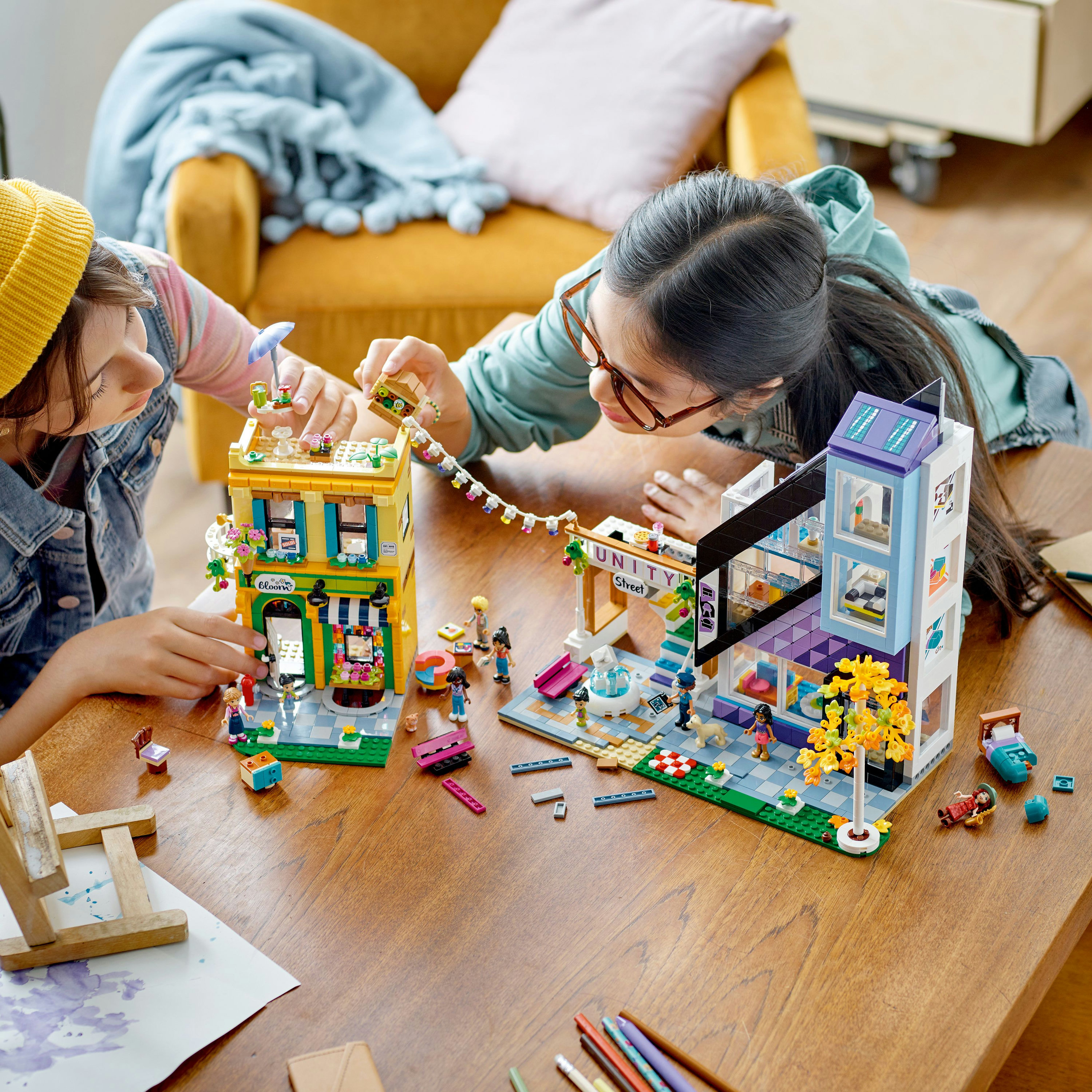 Dos niñas jugando con sets de Lego. Una construye una tienda, la otra un café. Una mesa de madera en primer plano.