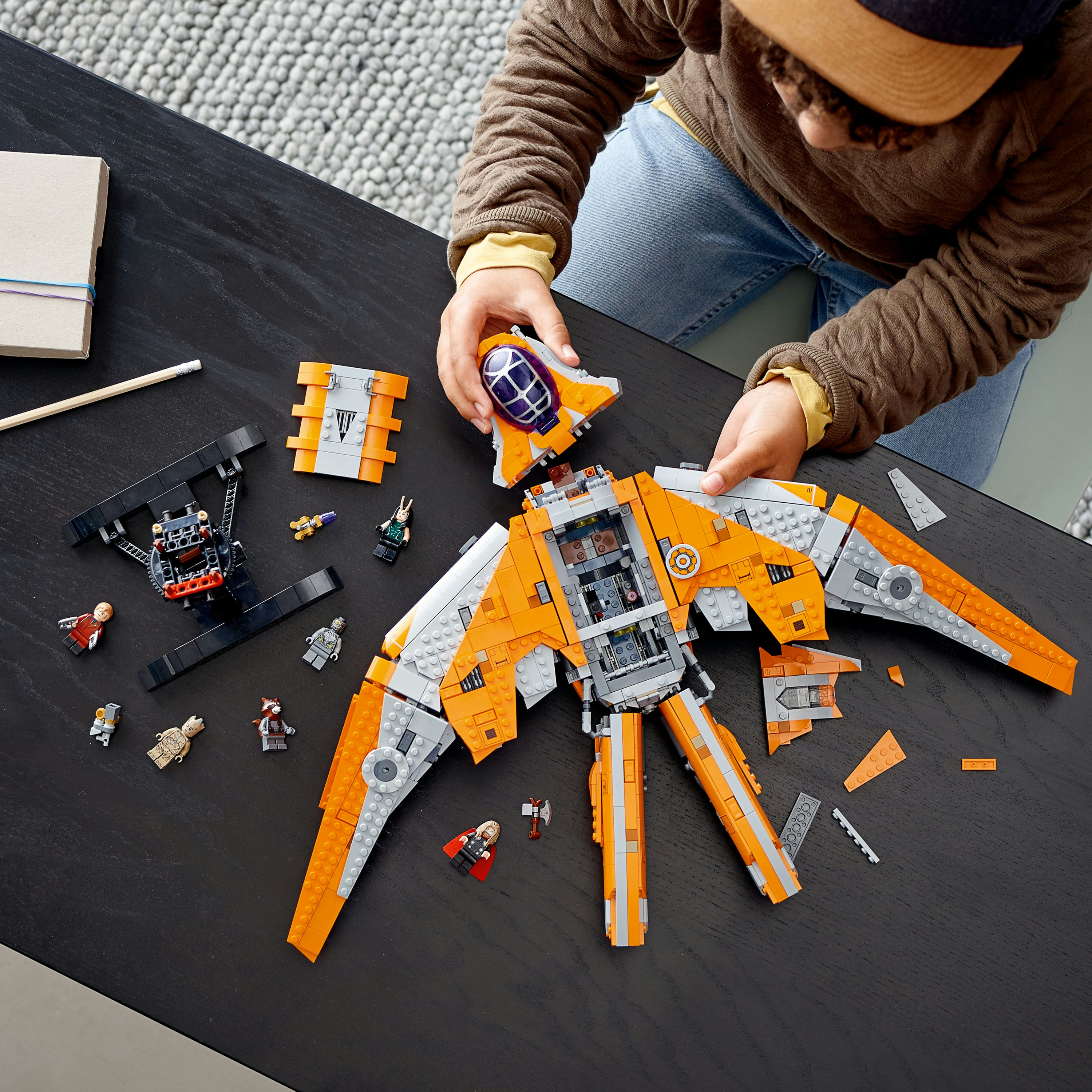 Kind baut LEGO Guardians of the Galaxy Schiff. Das Schiff ist orange und grau. Teile liegen auf Tisch.