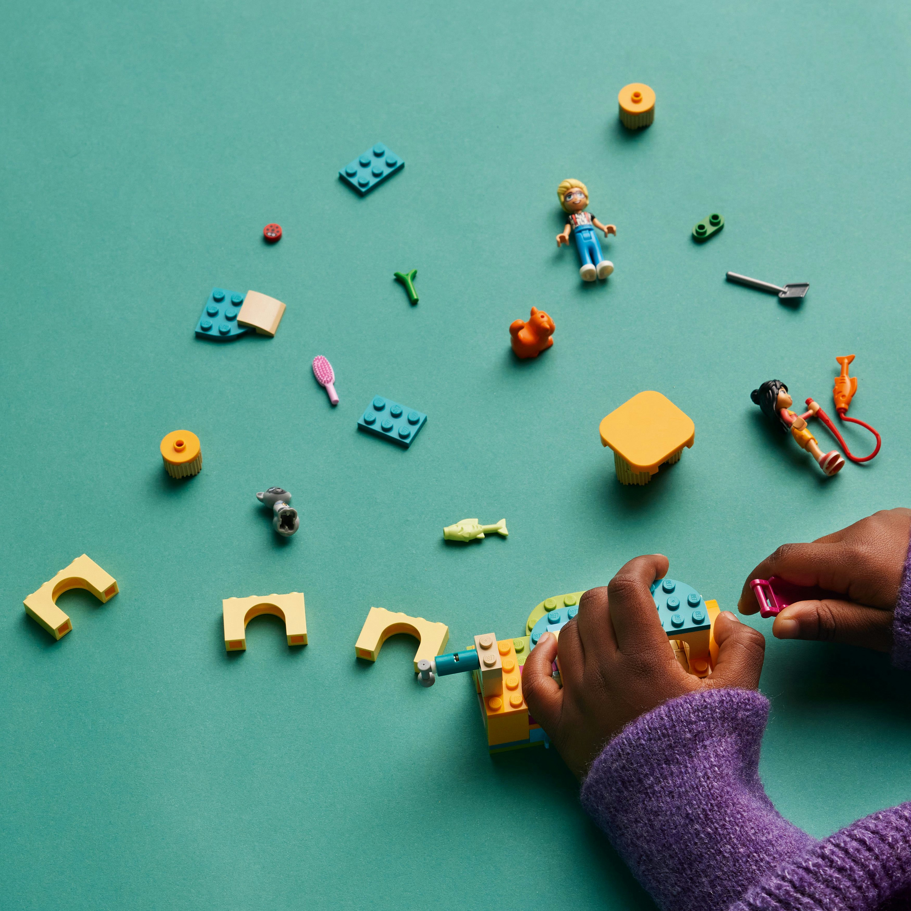 Hände, die LEGO-Steine zusammenbauen. Verschiedene LEGO-Teile und Figuren liegen auf grüner Oberfläche.