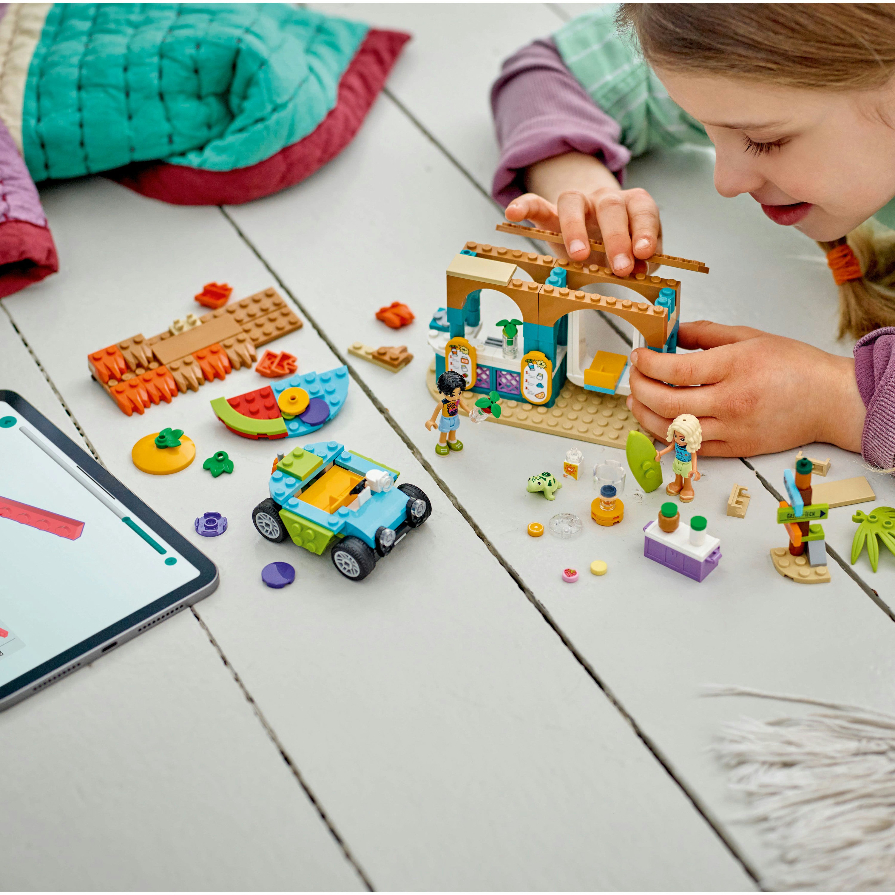 Dziewczynka buduje zestaw Lego na drewnianej podłodze. Zawiera samochód i sklep z kolorowymi elementami.
