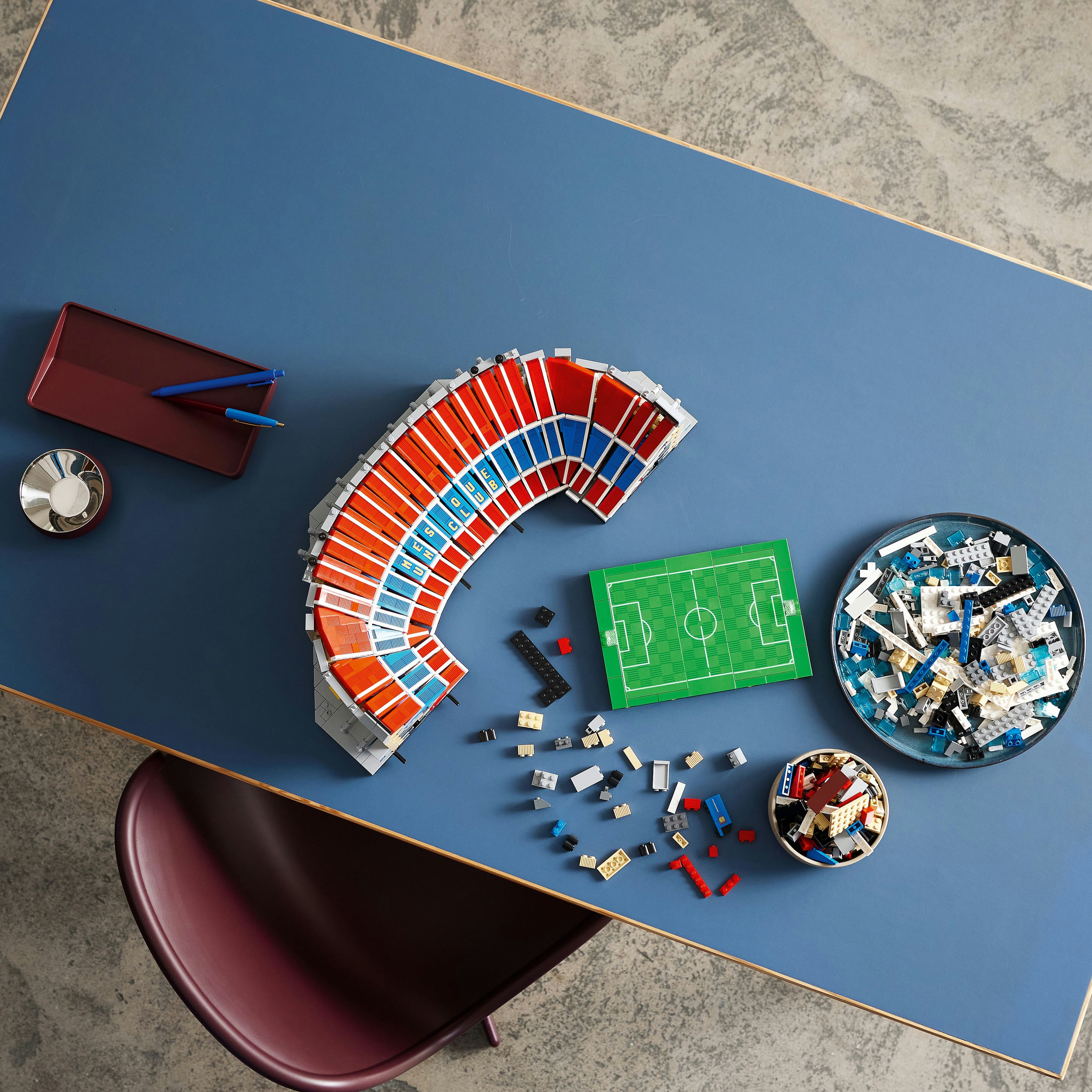 Zestaw Lego przedstawia model stadionu z boiskiem piłkarskim i rozrzuconymi klockami Lego na niebieskim stole.