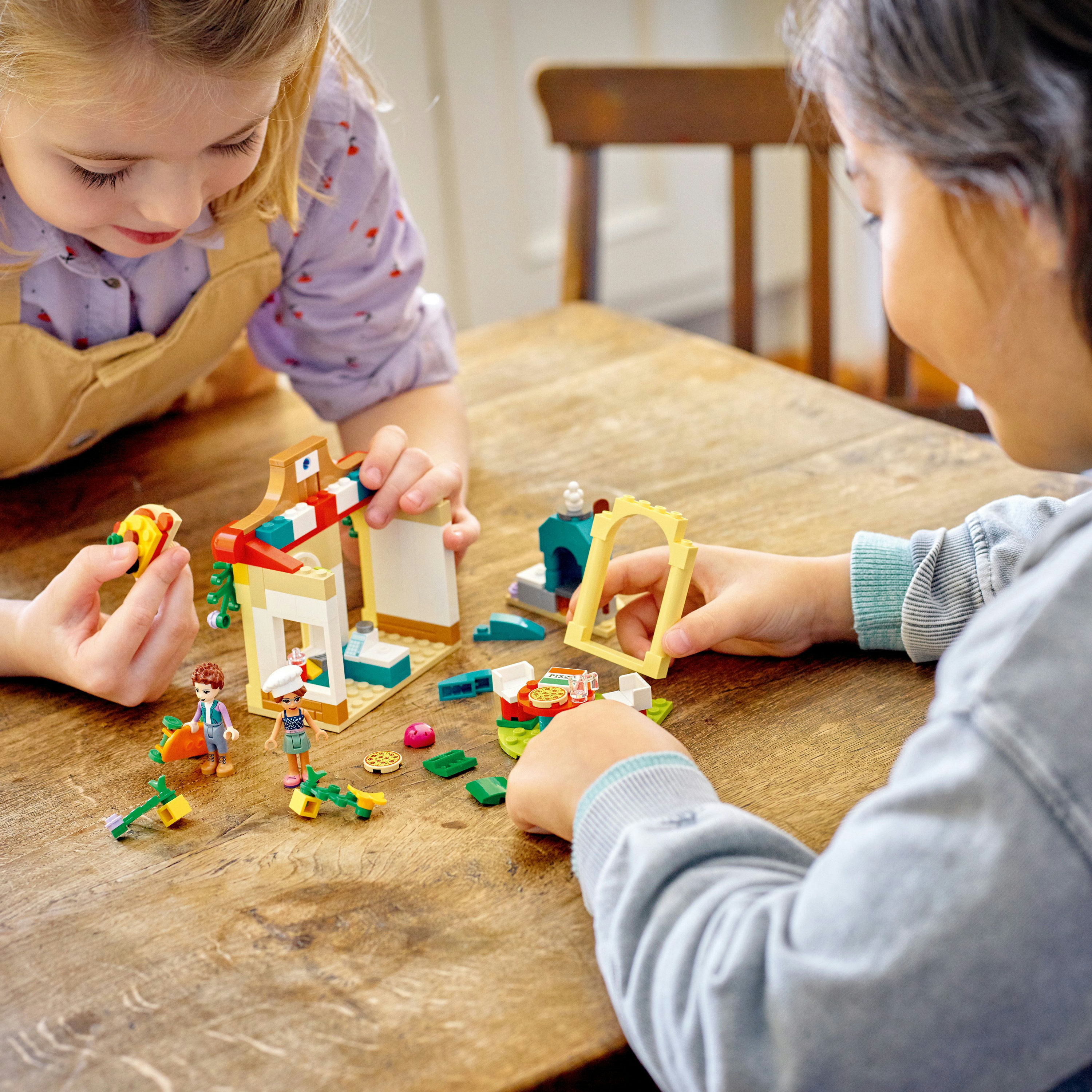 Zwei Kinder spielen mit Lego-Steinen auf einem Holztisch. Mehrere Lego-Elemente sind sichtbar.