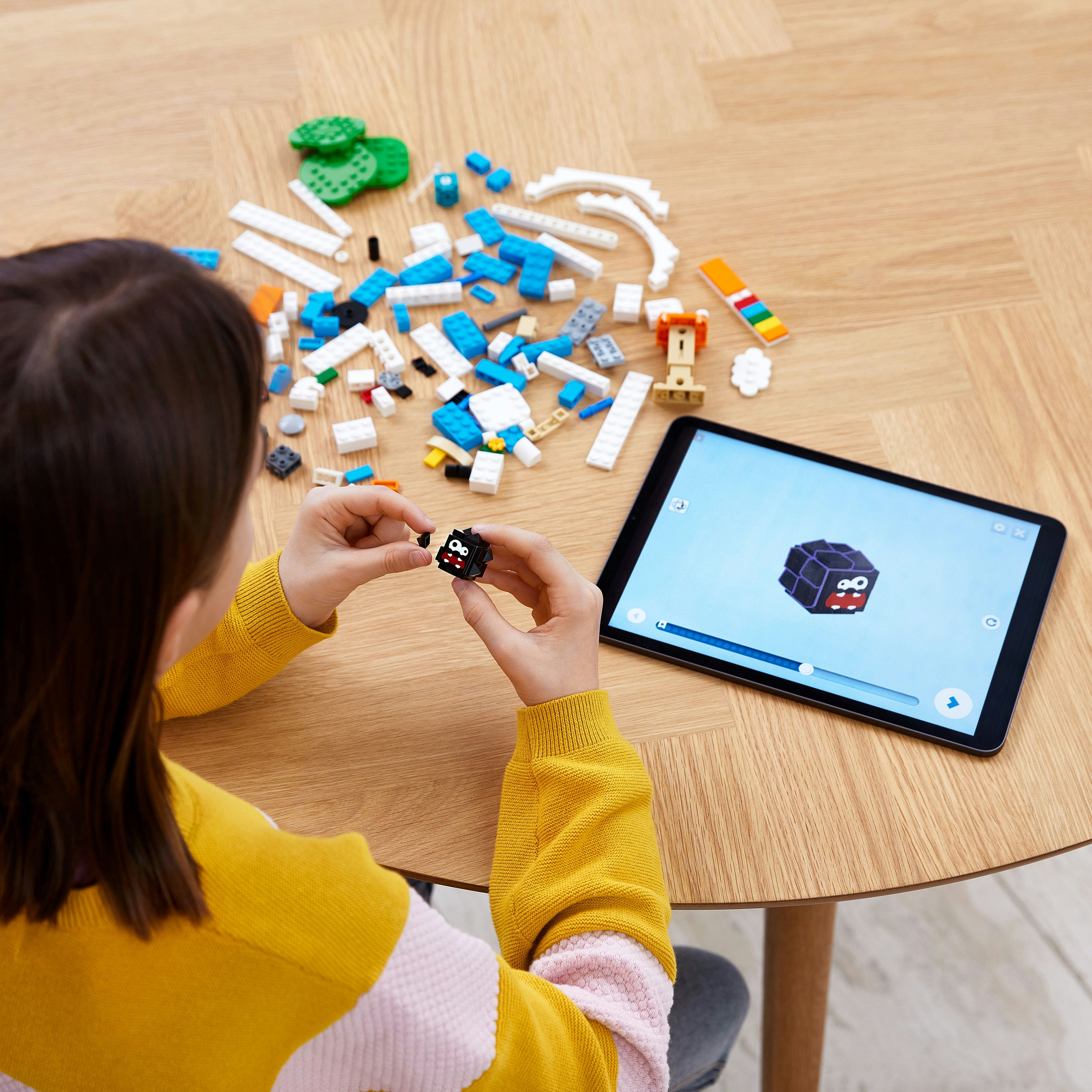 A child building a Lego block on a wooden table, looking at a tablet showing instructions for the build.
