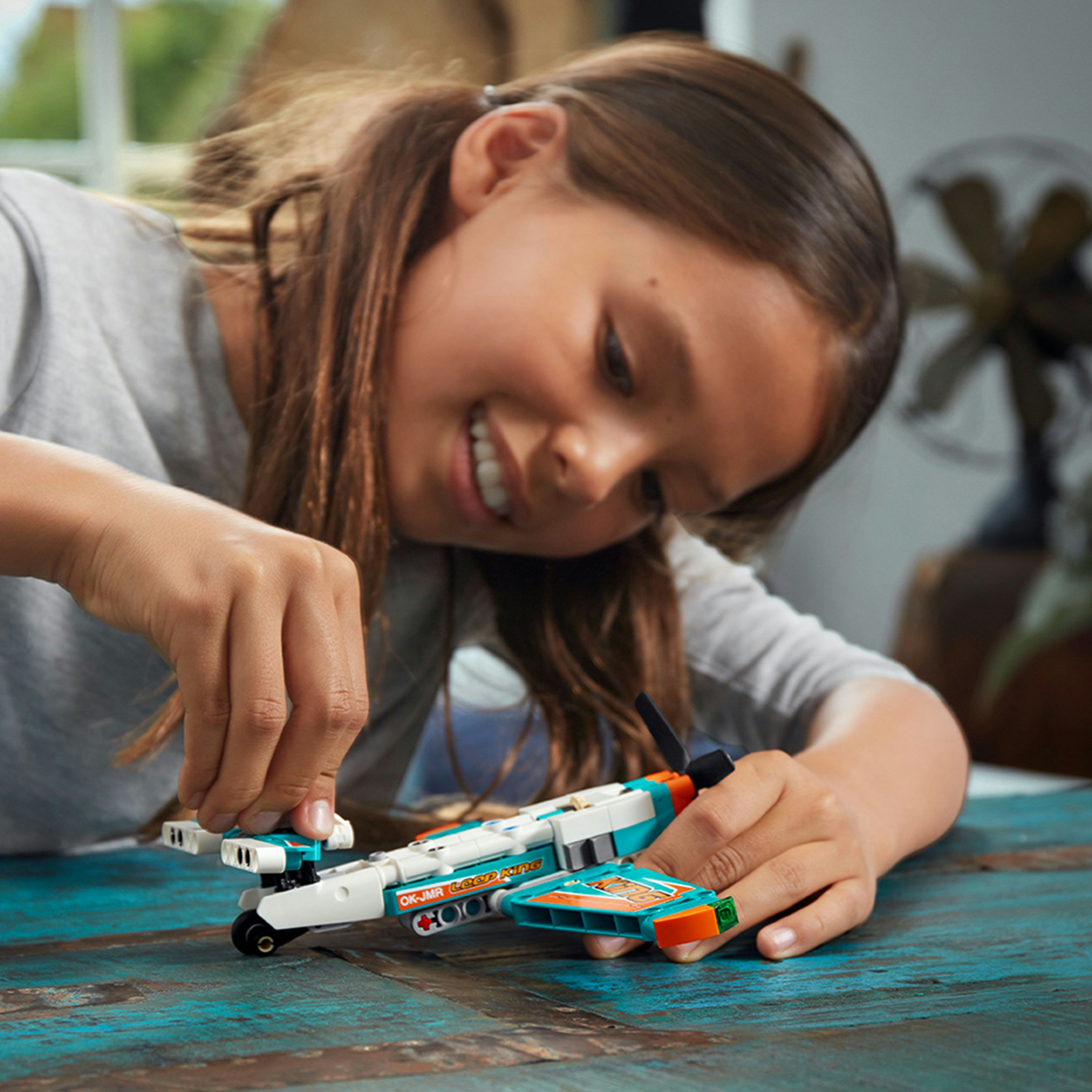 Ein junges Mädchen spielt mit einem Lego-Flugzeug auf einem Holztisch und lächelt in die Kamera.