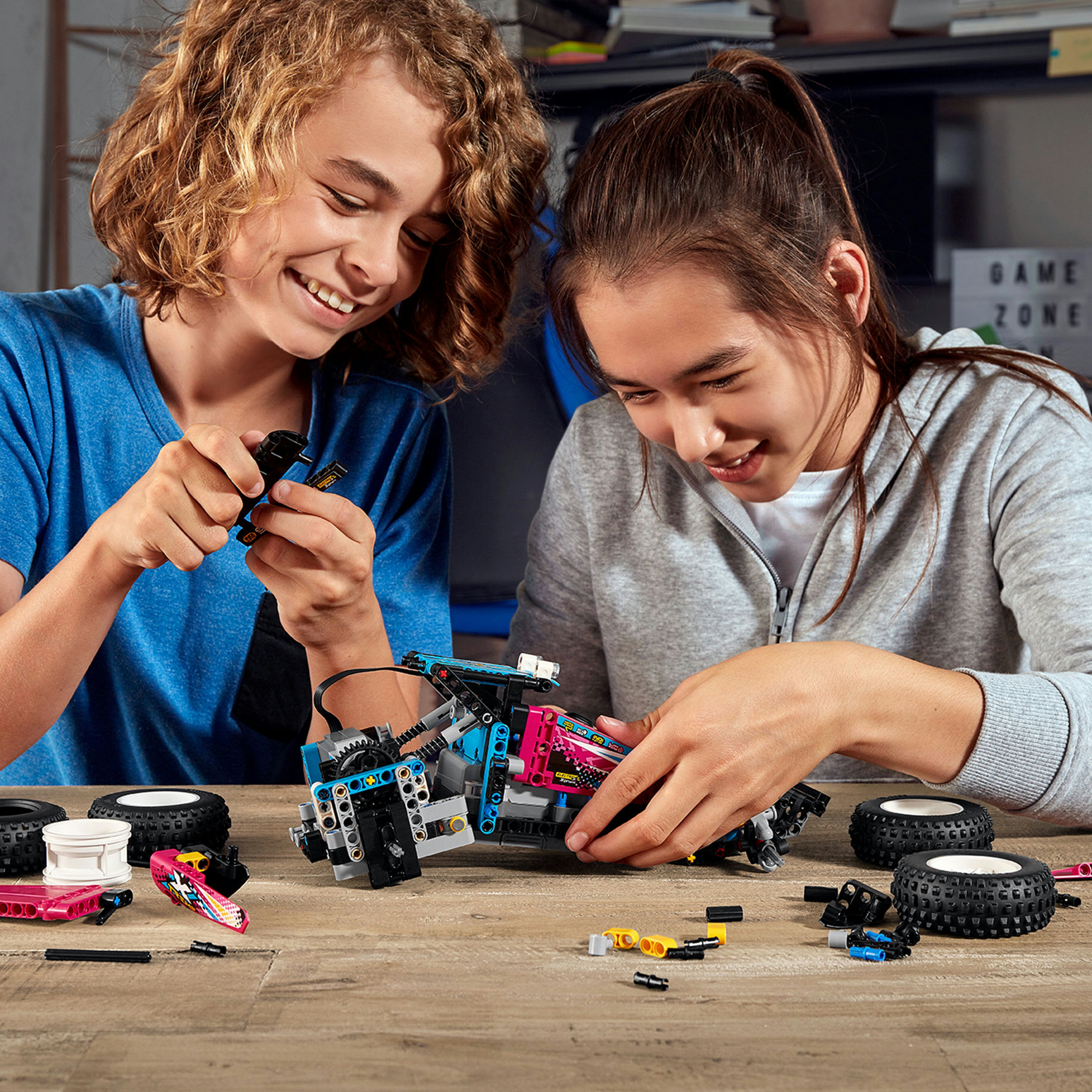 İki çocuk ahşap bir masada bir Lego Technic modeli topluyor. Çeşitli Lego parçaları etrafa dağılmış.