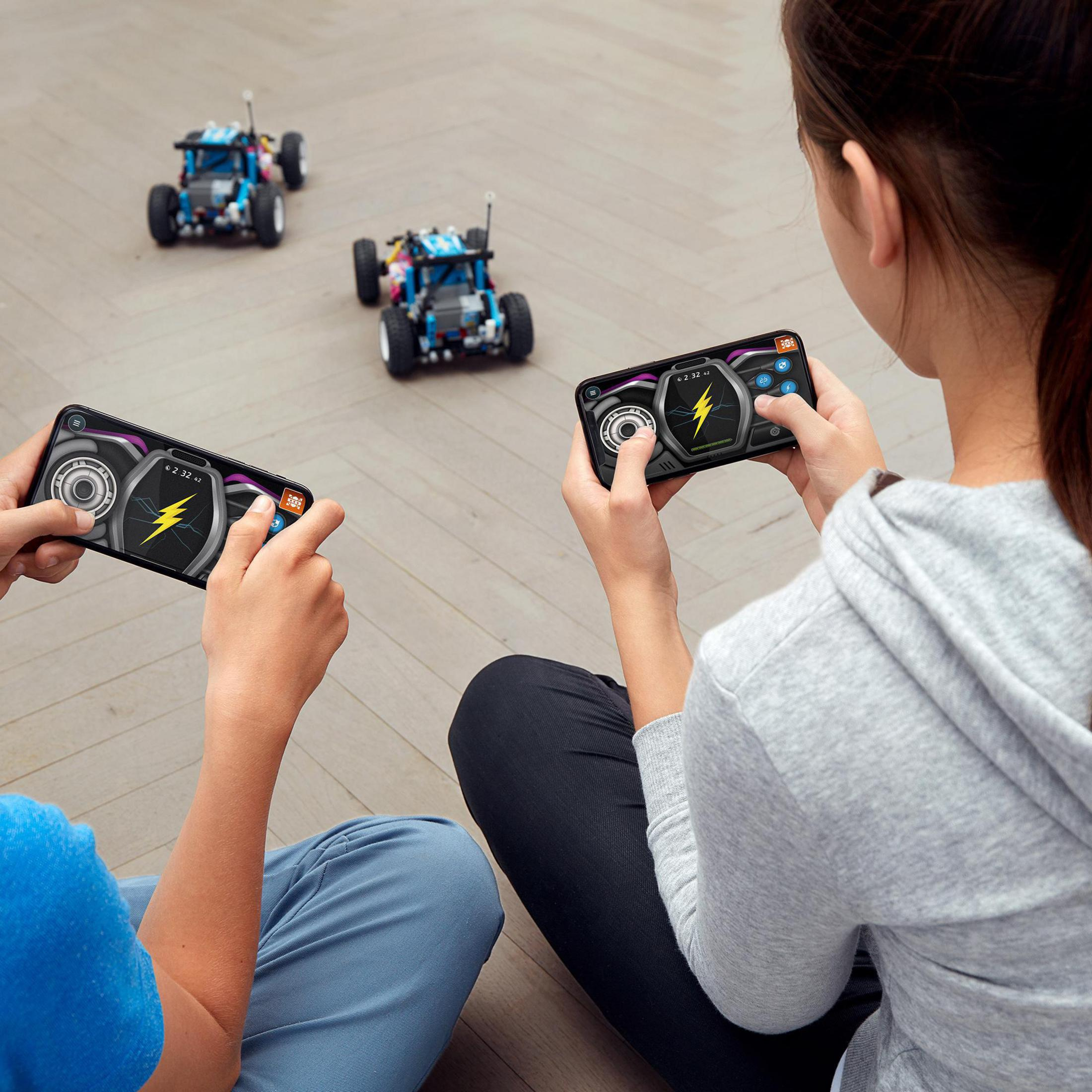İki kişi akıllı telefonlarla Lego arabalarını kontrol ediyor. Arka planda iki araba.