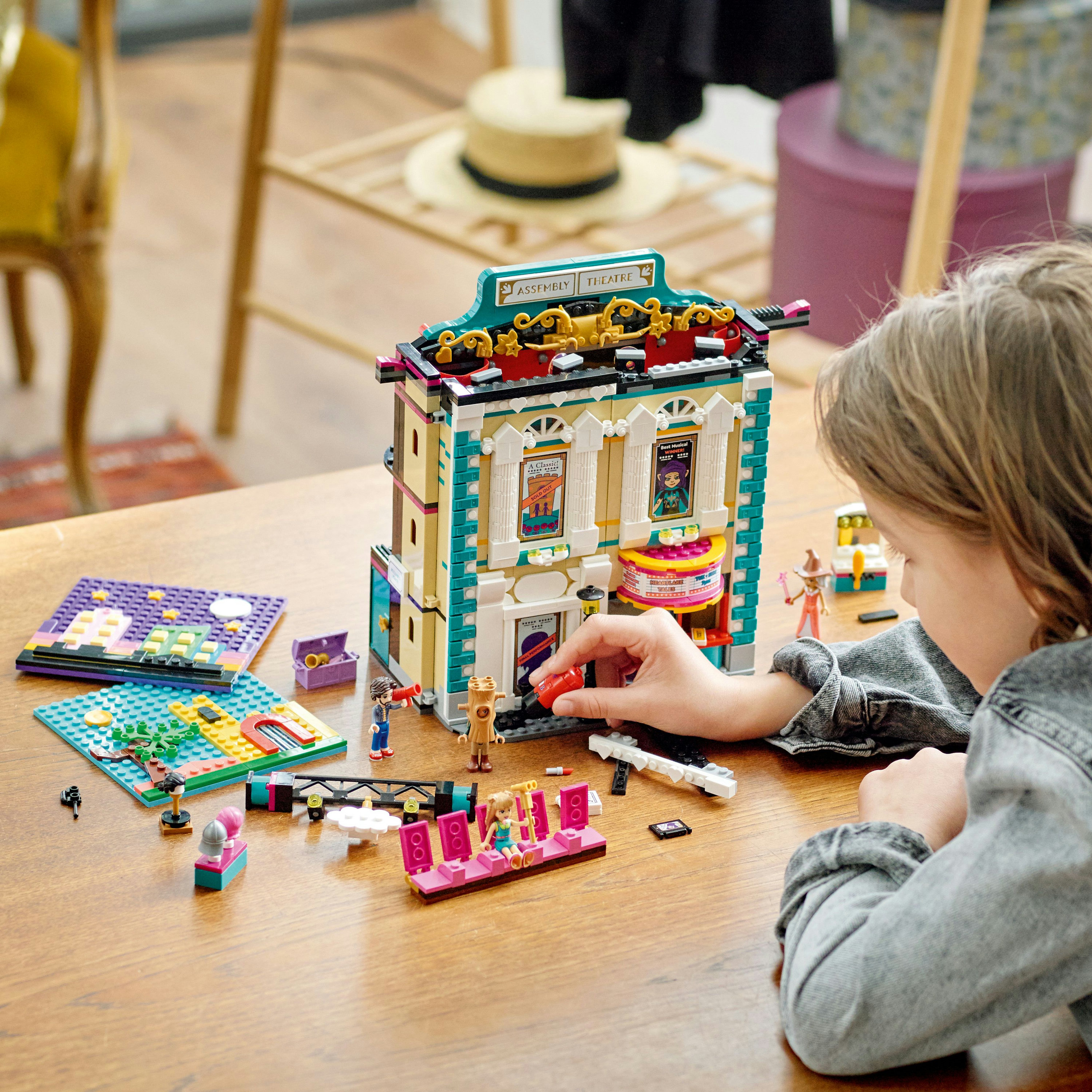 Ein Kind spielt mit einem Lego-Theater-Set auf einem Holztisch. Lego-Figuren und Requisiten sind verstreut.