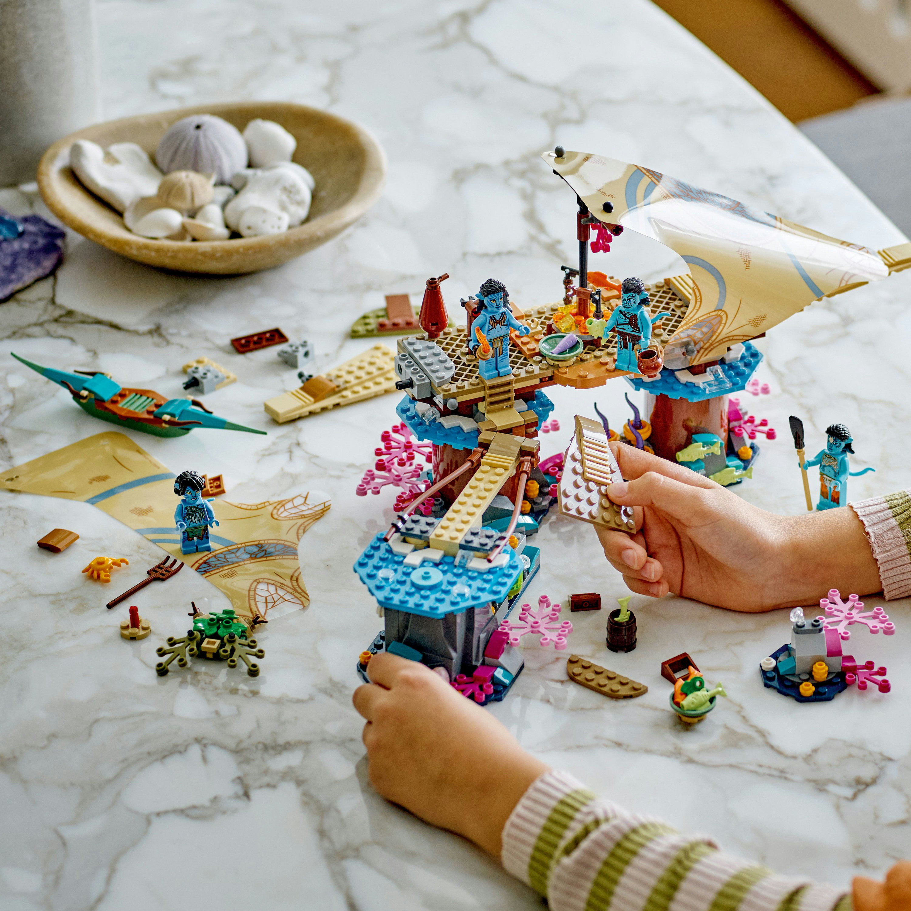 Set LEGO Avatar con figuras, estructuras y manos construyendo la escena sobre una superficie de mármol.