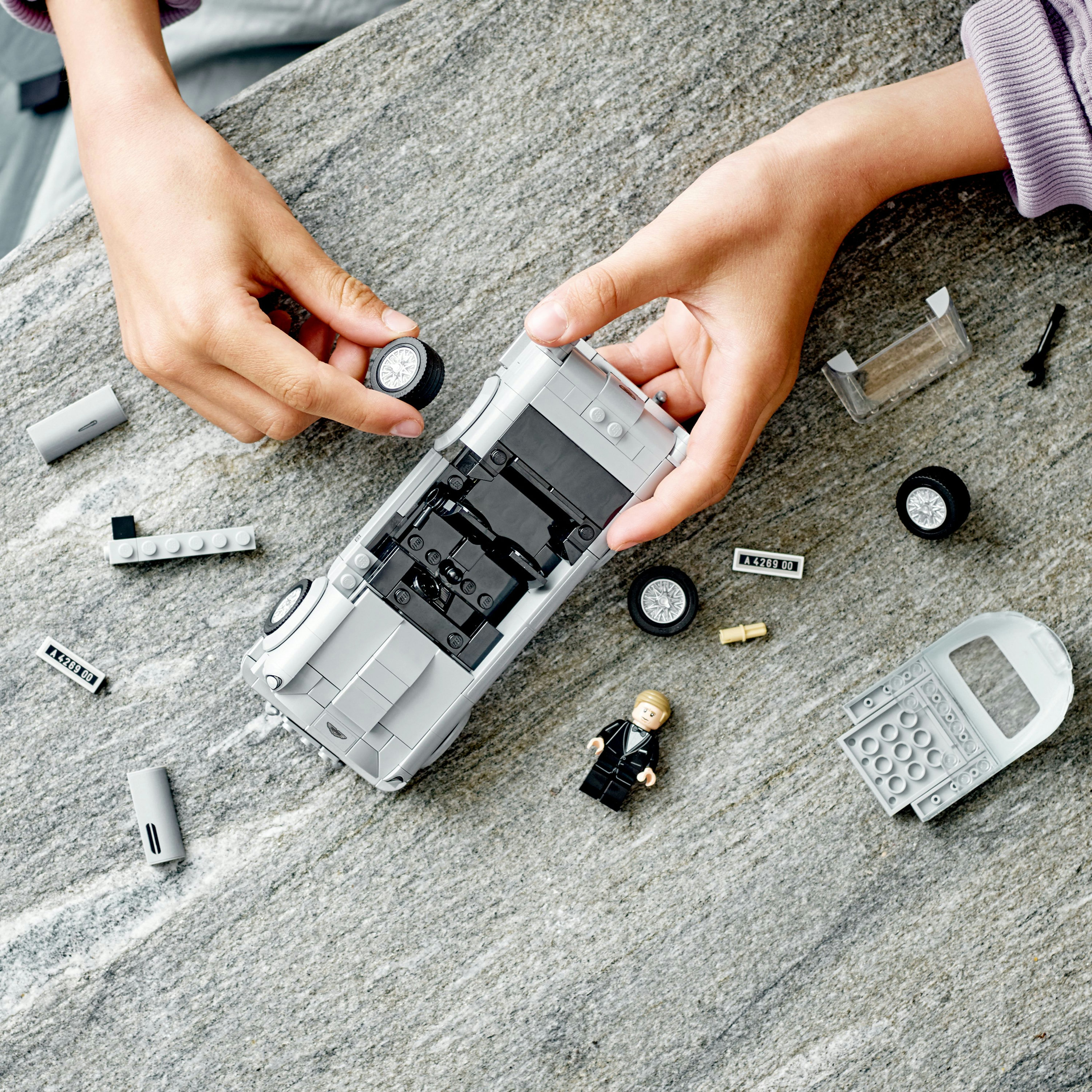 Hände montieren ein graues Lego-Auto. Verschiedene Teile sind sichtbar, einschließlich Räder und eine kleine Figur. Draufsicht.